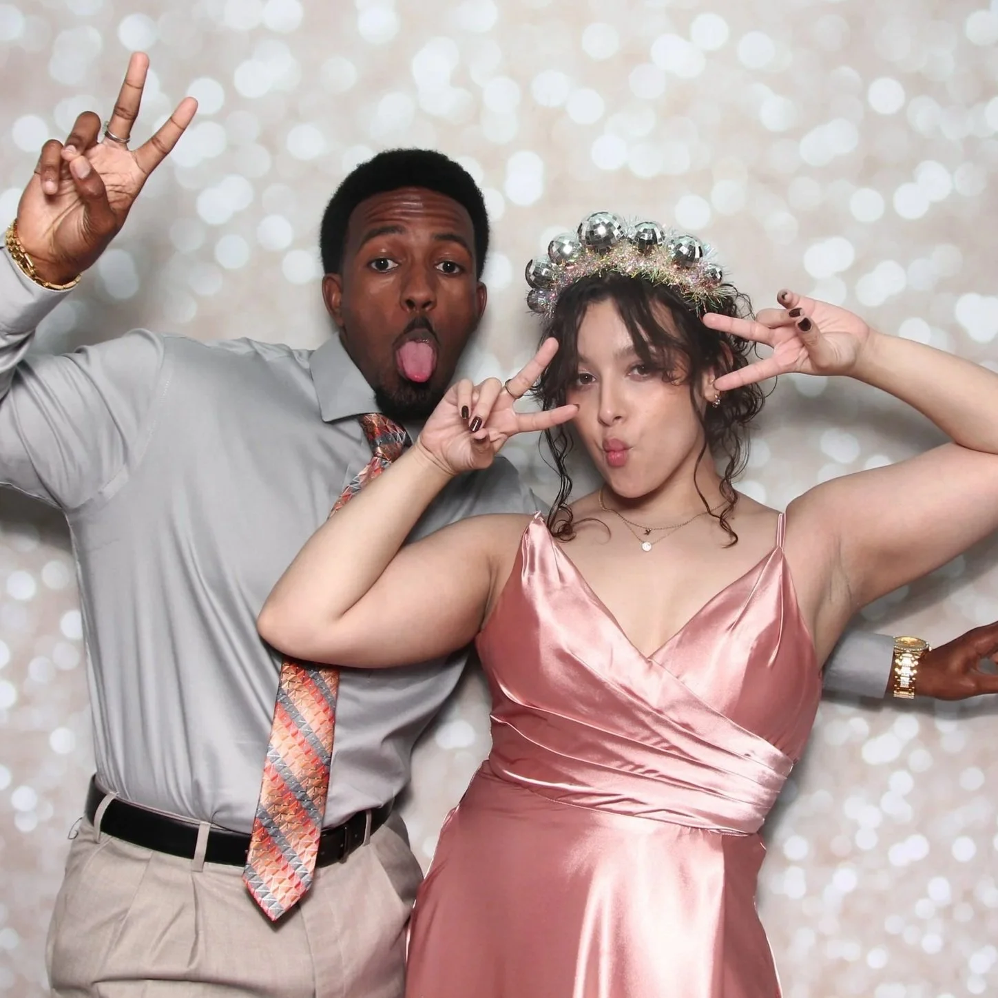 Two people in party attire making playful poses, with a sparkly background. The woman wears a pink satin dress and a festive headpiece, and the man wears a light gray shirt with a patterned tie.