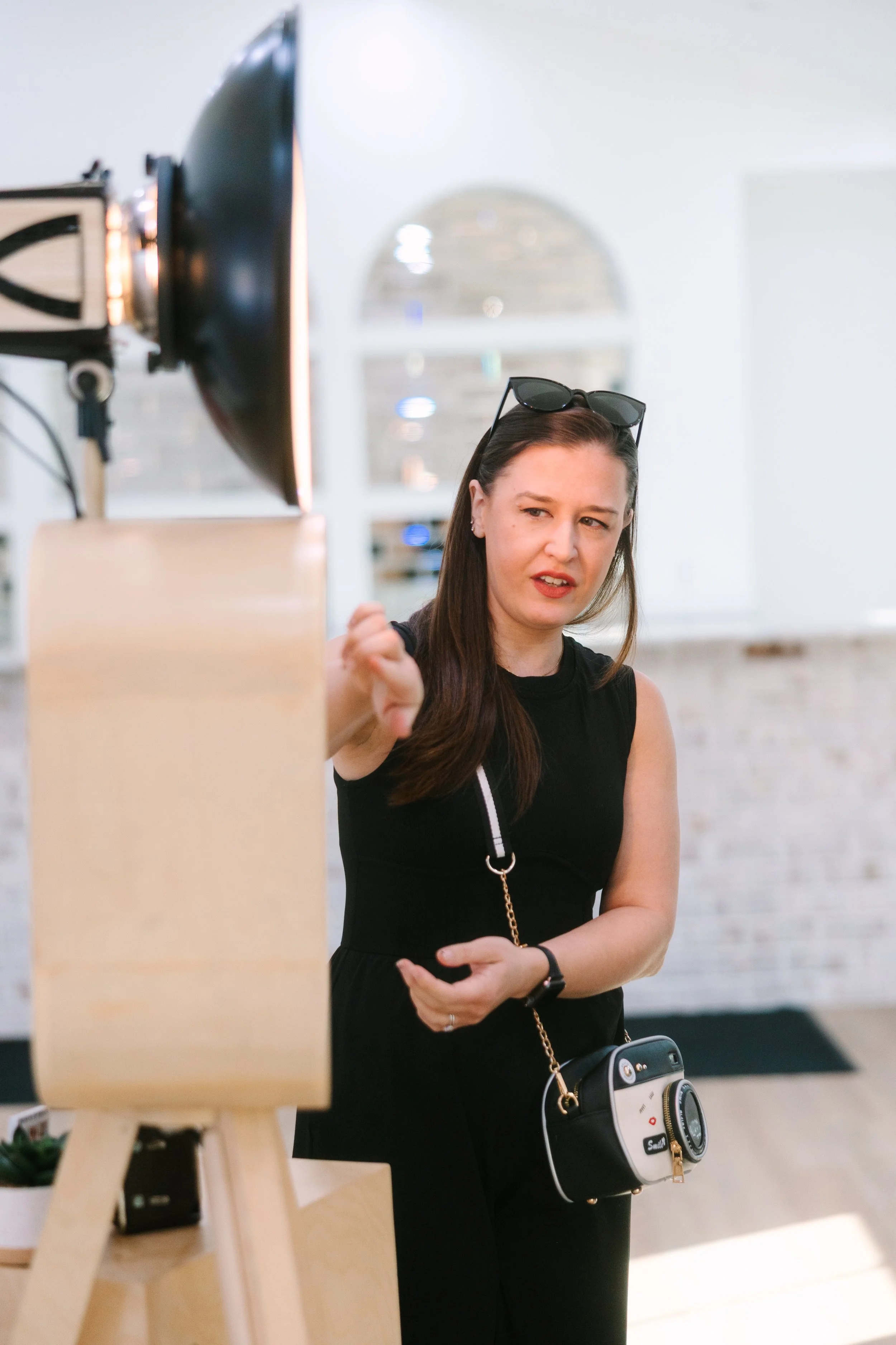 Woman with sunglasses on her head looks confused or skeptical while standing indoors near a photography studio setup with a large light and a wooden easel.