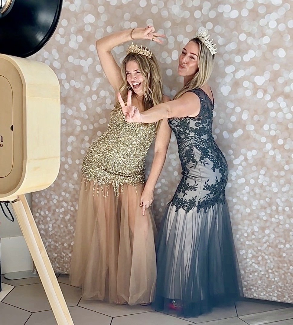 Two women wearing crowns and glamorous evening gowns taking a selfie together in front of a glittery photo booth backdrop.