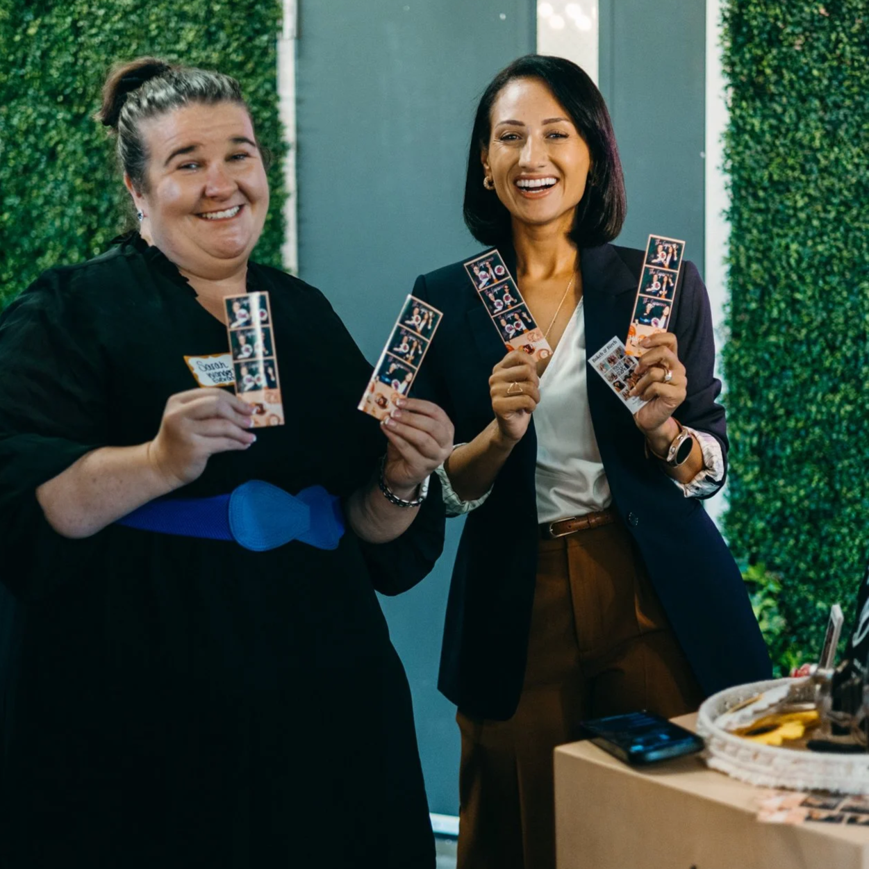 Two women smiling and holding photo booth strips at an indoor event with green foliage background.