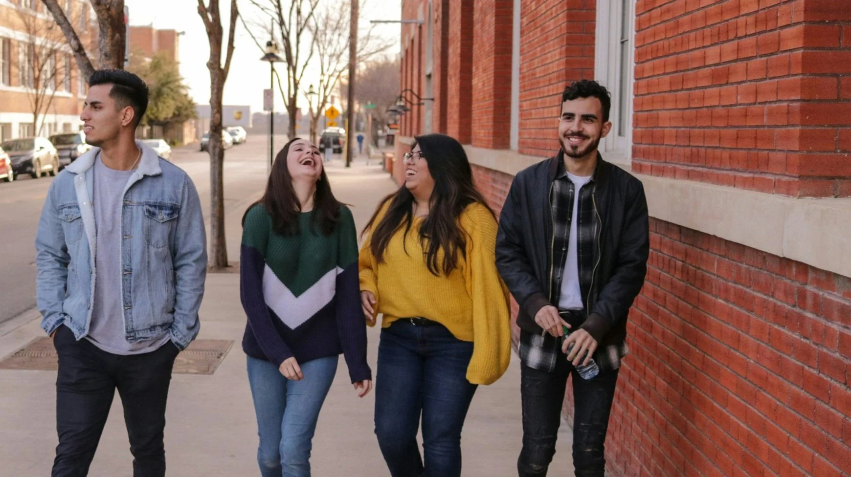 Two girls, wearing green and yellow sweaters, are laughing together and walking down a historic brick street.