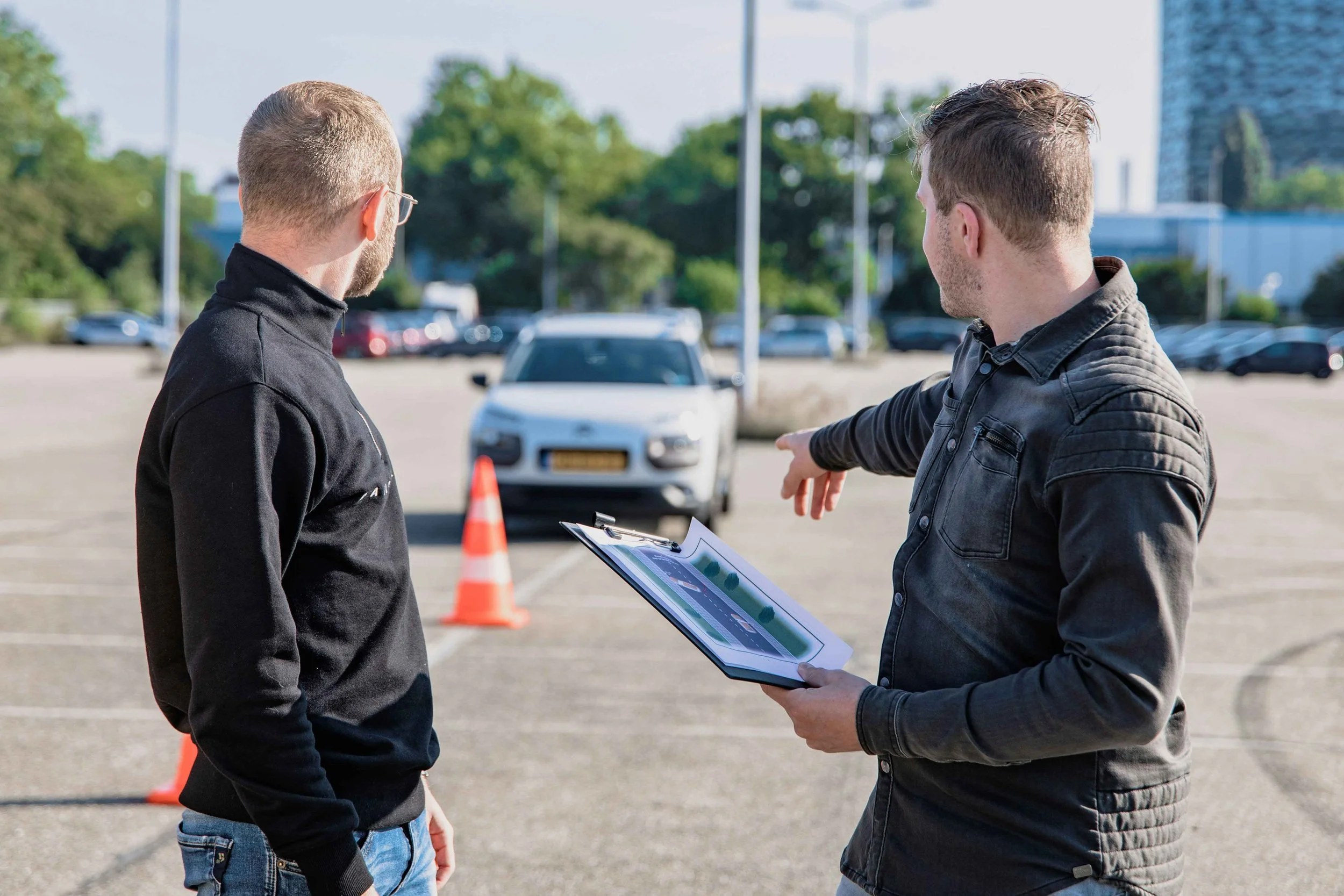 Two guys wearing black jackets are turning and looking at a car behind them. One man is holding a clipboard and pointing to the car. In front of the car is an orange cone, and behind are other cars parked near trees in the background.