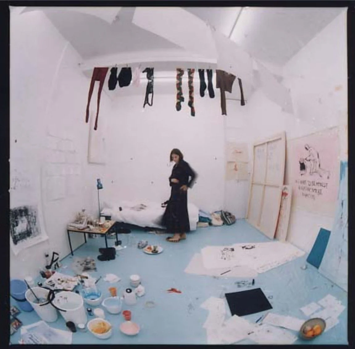 Fish-eye view of Tracey Emin's studio installation Life Model Goes Mad, 1996, showing a cluttered blue floor, hanging garments, canvases leaning against white walls, and a figure in dark clothing mid-movement