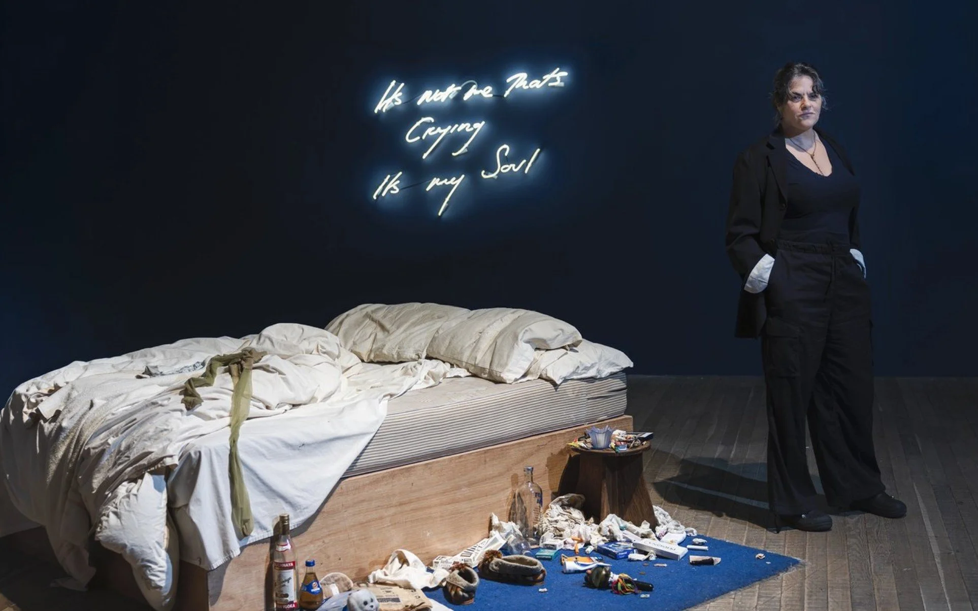 Tracey Emin stands beside her installation My Bed at Tate Modern, dressed in black, with neon text reading "It's not me that's crying, it's my soul" glowing on the dark wall behind her