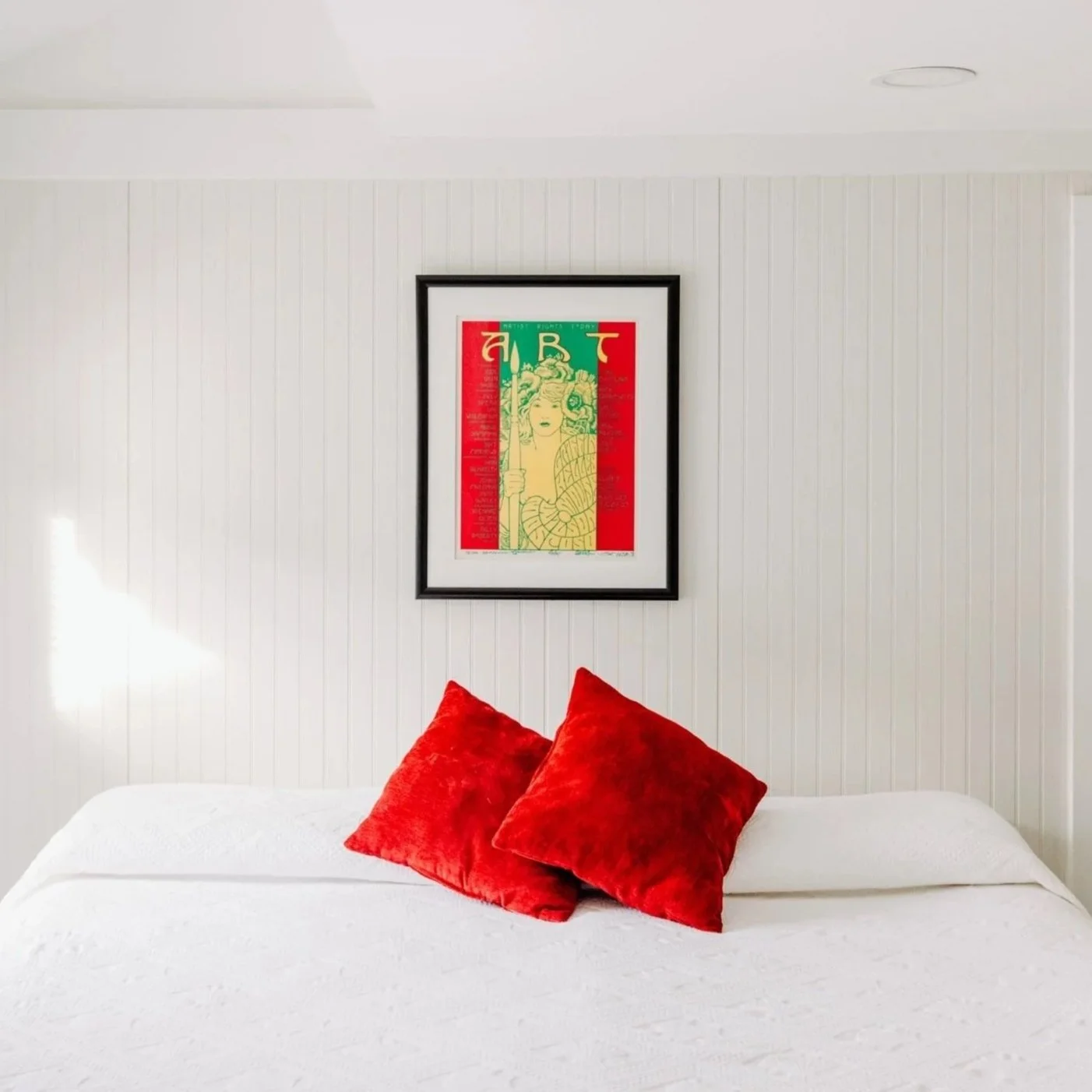 White bed with two red velvet pillows against a white paneled wall, with framed colorful artwork hanging above.