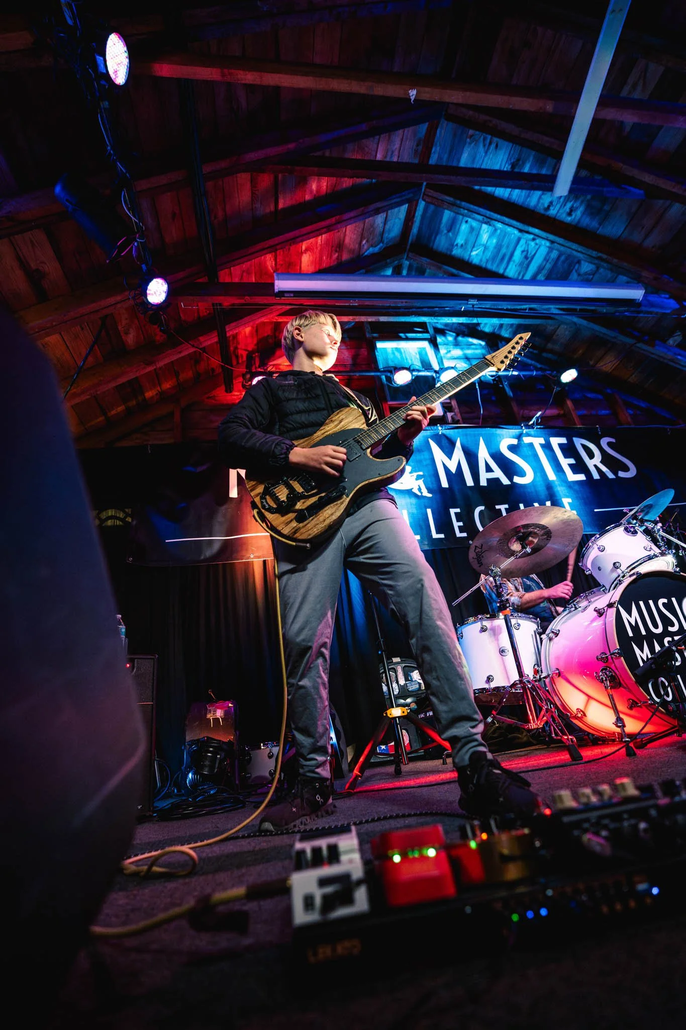 A band performing on stage with a guitarist in the foreground and a drummer in the background, under colorful stage lighting.