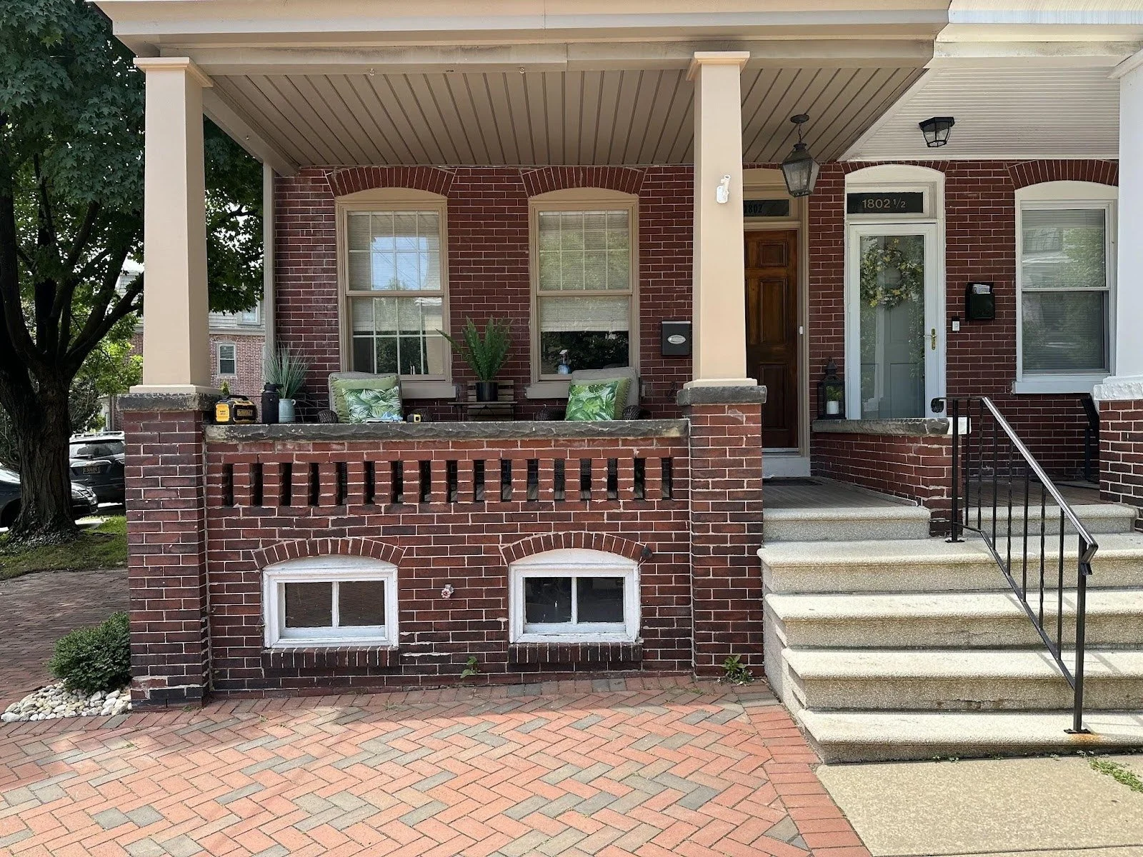 Front porch of a brick house with steps leading up to the entrance, two hanging windows below, and a small patio area with potted plants and chairs.
