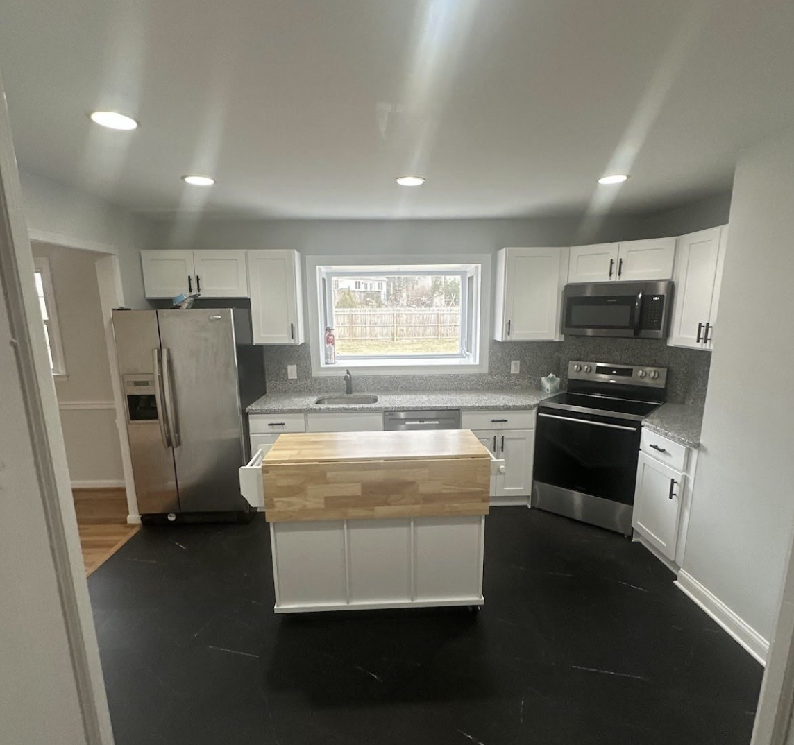 Modern kitchen with white cabinets, stainless steel refrigerator, black microwave and oven, gray granite countertops, a small kitchen island, and a window overlooking a backyard with a wooden fence.