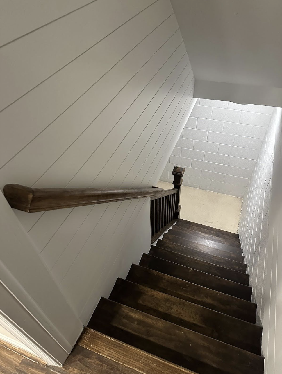 A staircase with dark wooden steps and a wooden handrail on the left side, leading down to a basement area with concrete floor and concrete block walls painted white.