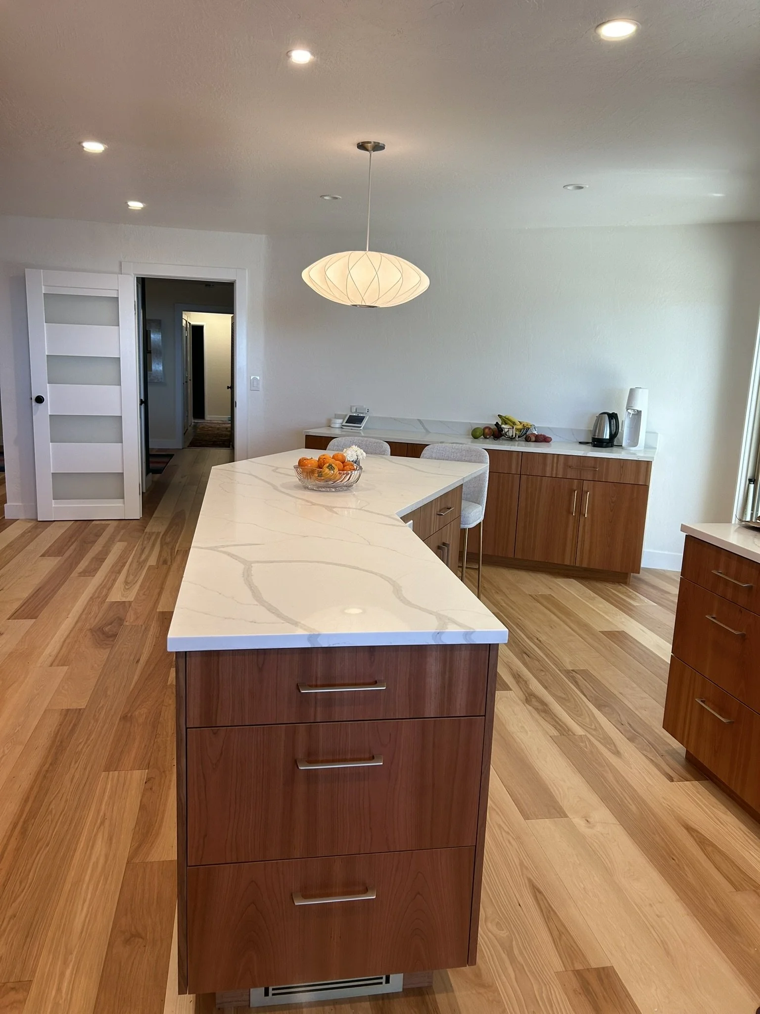 Modern kitchen with a white marble island, wooden cabinetry, fruit bowl, and appliances on a white countertop. Recessed lighting and hardwood floors are visible.