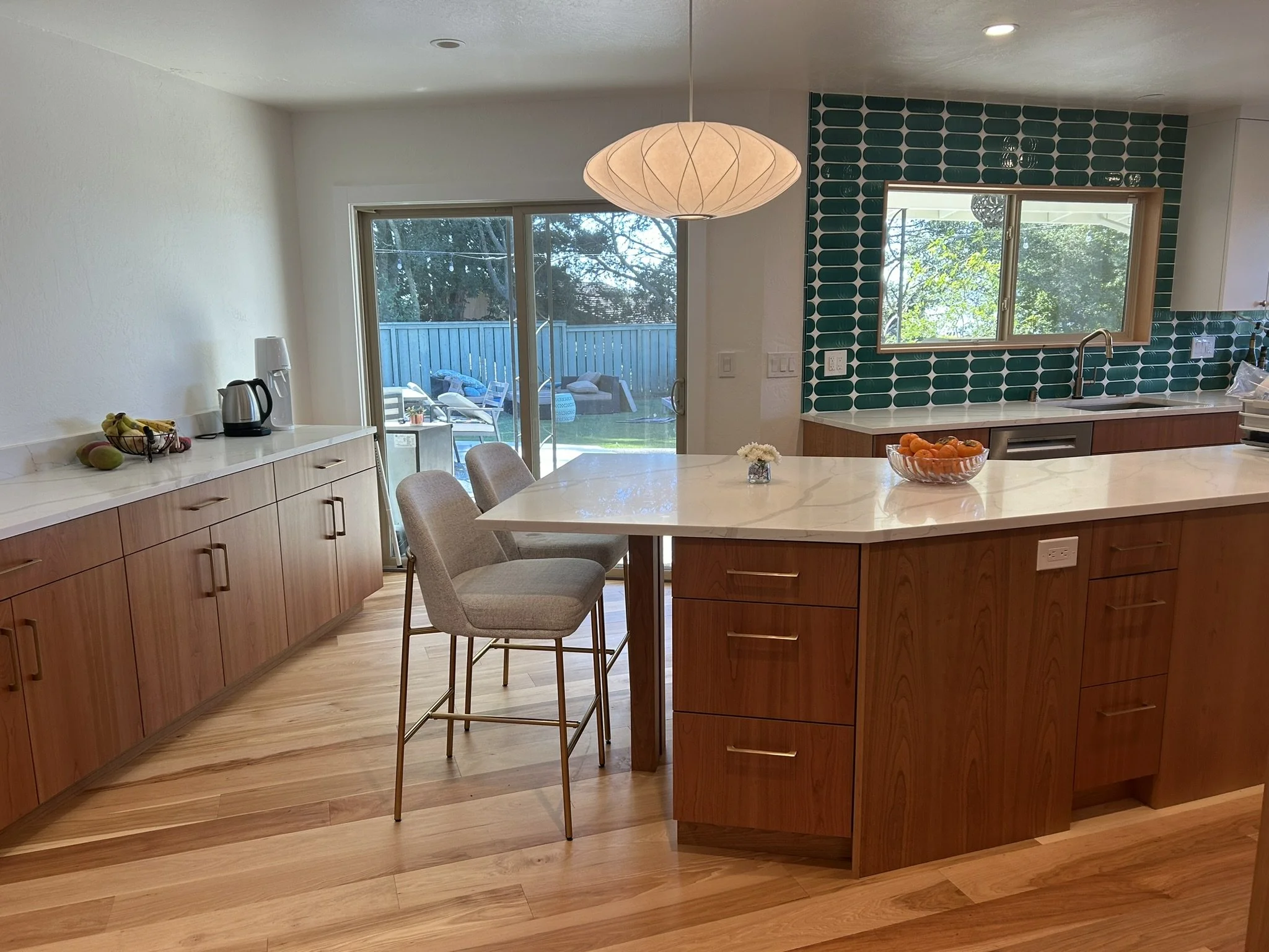 A modern kitchen with a marble island, wooden cabinets, green tiled backsplash, and sliding glass door leading to an outdoor patio with chairs and a blue fence.
