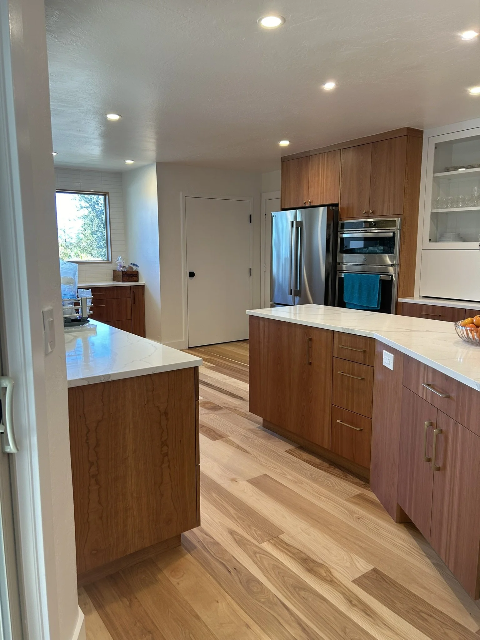 Modern kitchen with wooden cabinets, stainless steel appliances, hardwood floors, white countertops, and a small window with outside greenery.