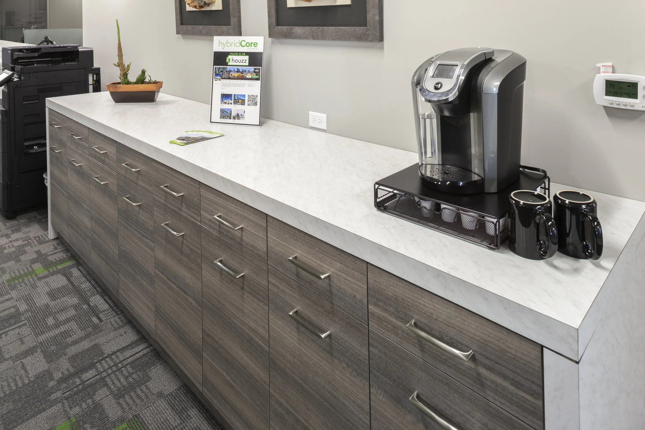 A modern office kitchenette area with a white countertop, wooden cabinet drawers, a black coffee maker, a small plant, and a wall-mounted thermostat. There is a sign on the countertop with information and a QR code. The floor has gray patterned carpet.