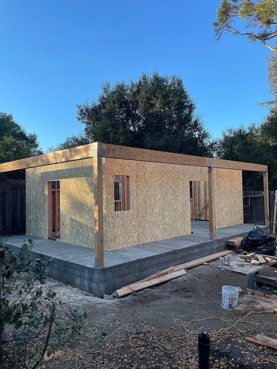 A house under construction with plywood walls, wooden framing, and a concrete foundation, in a backyard with trees and tools around.