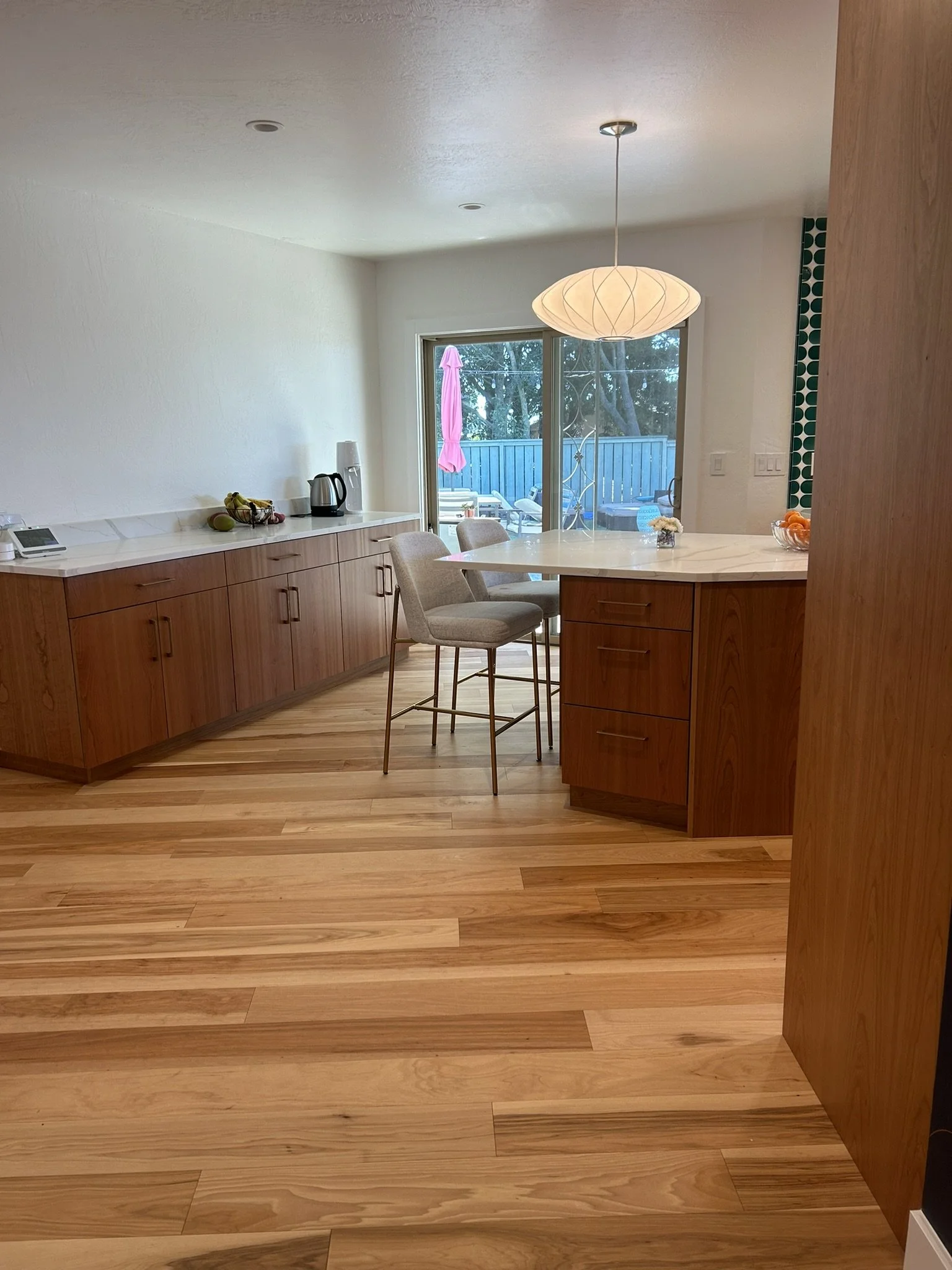 A modern kitchen with a wooden floor, white walls, a marble countertop, and a view of an outdoor patio with a blue fence, patio furniture, a pink umbrella, and trees through sliding glass doors.
