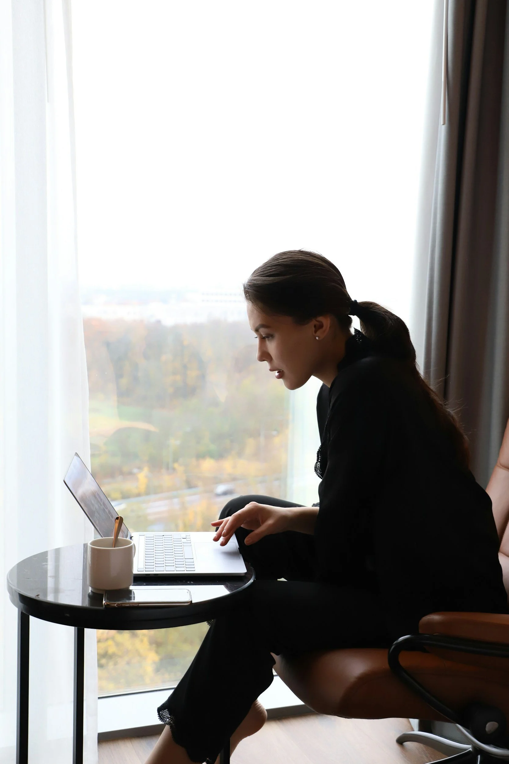 A woman sitting in a chair at a small round table by a large window, working on a laptop.