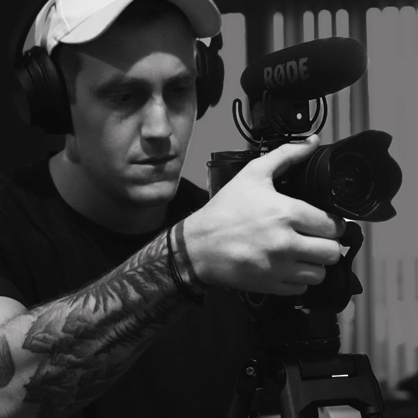 A young man wearing a cap and headphones, operating a professional video camera on a tripod, with a Rode microphone attached, in a studio or recording space.