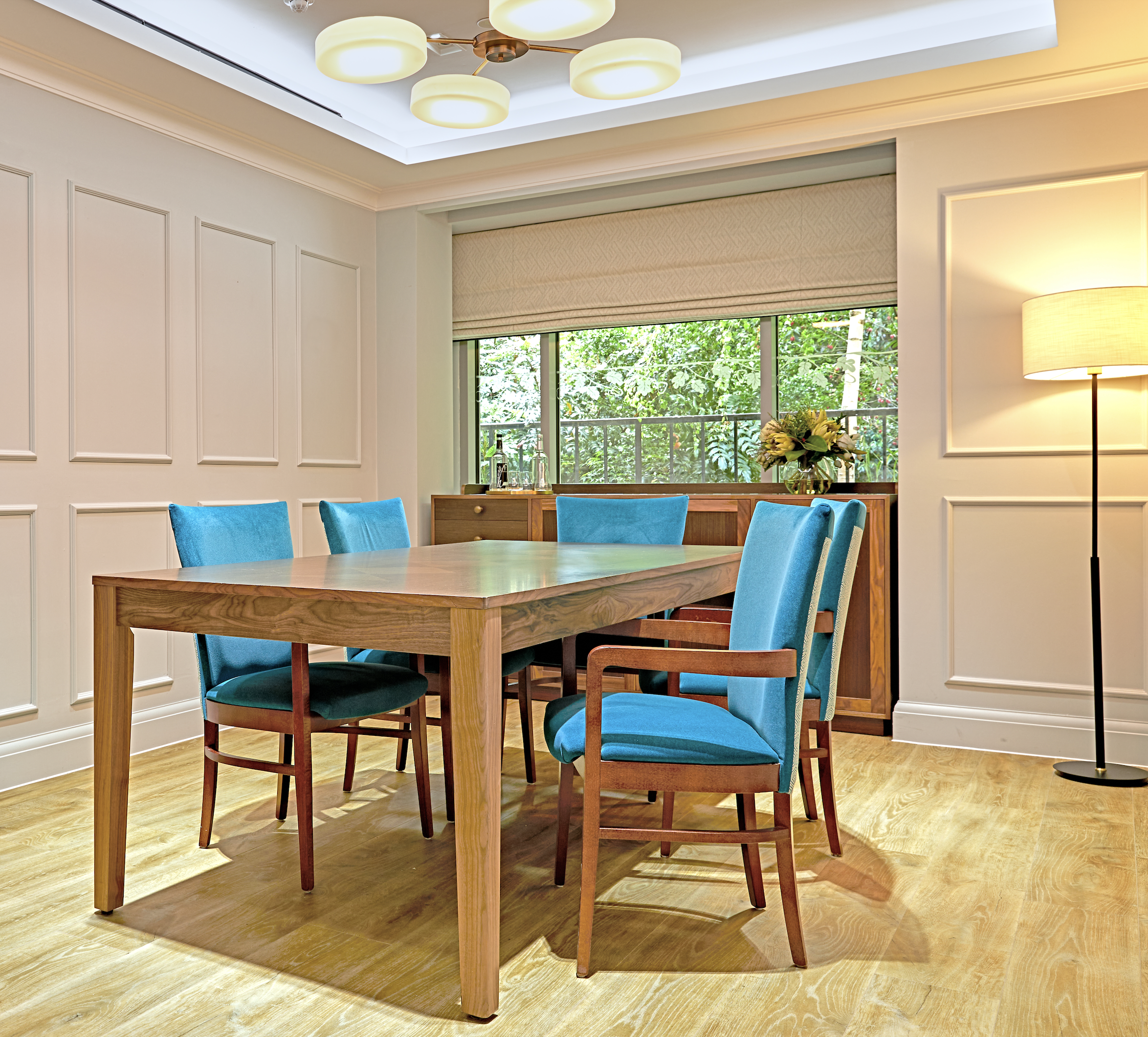 A dining room with a wooden table and six blue upholstered chairs. There's a large window with a view of greenery outside. A flower arrangement is on a wooden sideboard, and a floor lamp stands in the corner. The ceiling has modern circular light fix