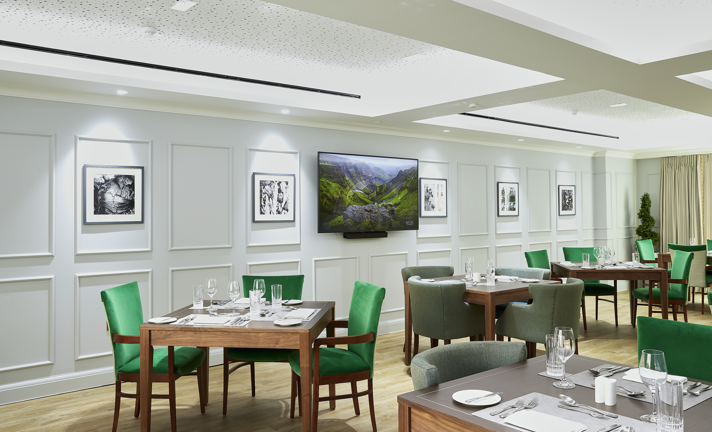 Elegant restaurant with white paneled walls and green upholstered chairs, set with glassware and silverware, and a large flat-screen television on the wall.