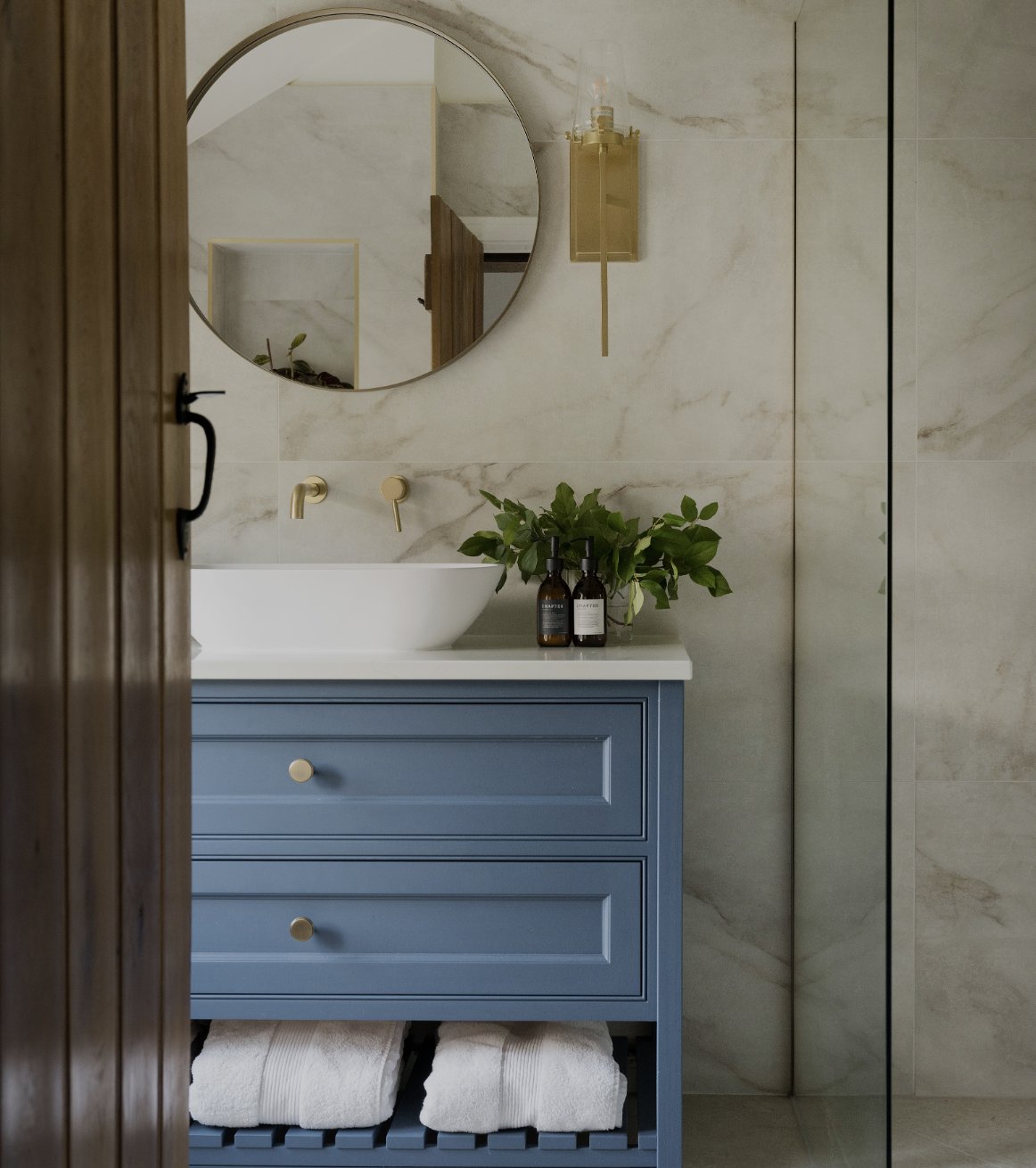 Harrogate-style bathroom designed by StyleSmith Interiors with blue vanity, white vessel sink, gold faucet, round mirror, marble walls, and glass shower enclosure.