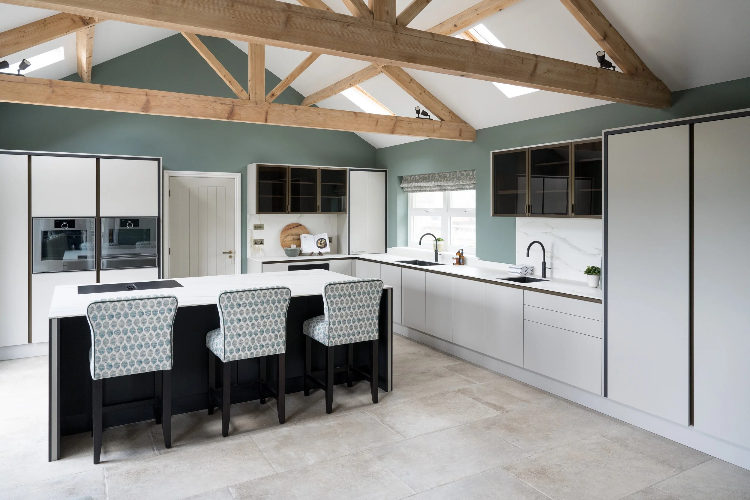 Open-plan kitchen and living space by Stylesmith Interiors with exposed oak beams, white cabinetry, black accents, and a contemporary island