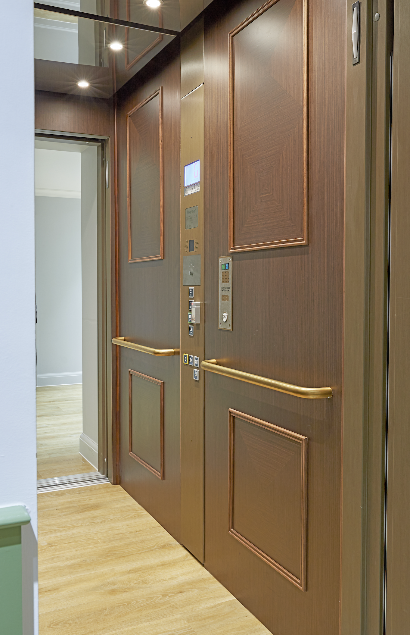 Interior view of a wooden elevator with closed doors, control panel with buttons and a display screen, located in a building with hardwood flooring.