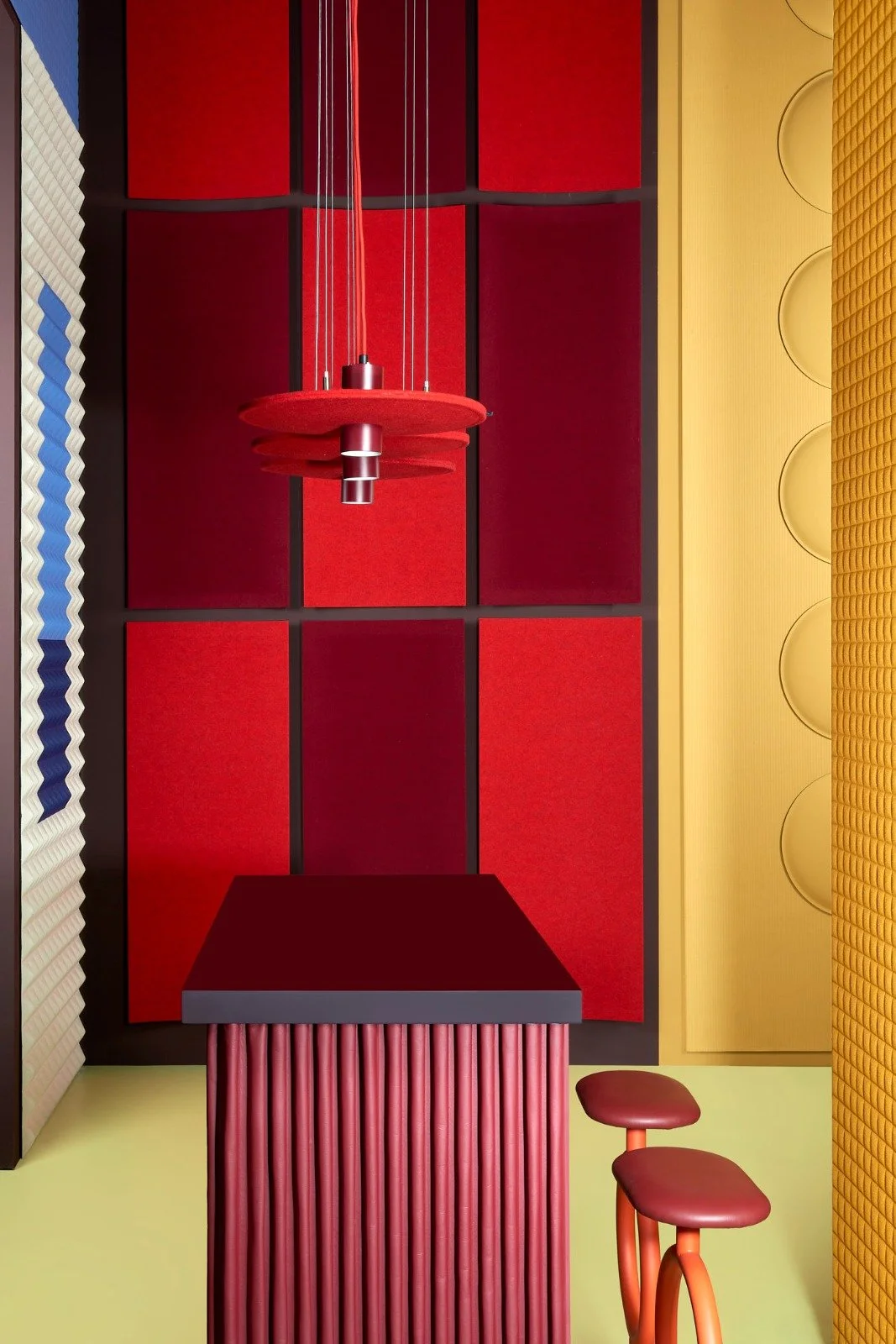 Modern interior with red and yellow geometric design, hanging red pendant light, red ribbed pillar column, and two red stools.