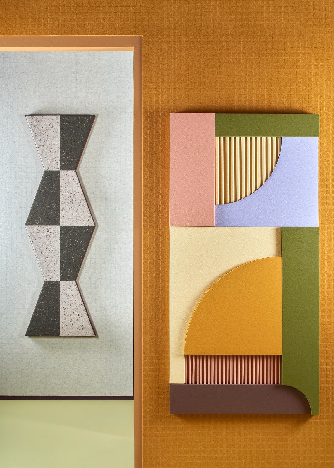 Colorful abstract wall art featuring geometric shapes, including a pink rectangle, blue quarter-circle, yellow quarter-circle, green rectangle, and pink ribbed rectangle, displayed on an orange textured wall.