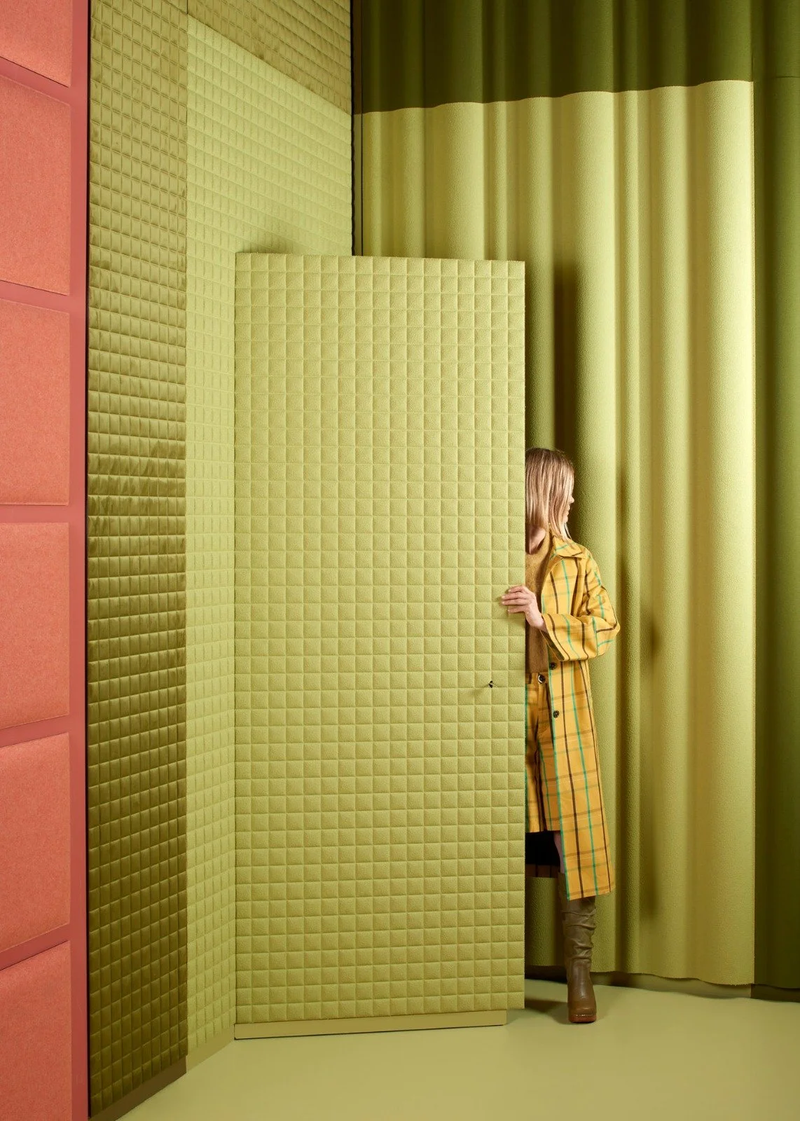 A woman in a yellow plaid trench coat stands behind a green padded foldable partition, peering out from behind it. The scene features green curtains, a green padded wall, and some pink padded panels on the left side.