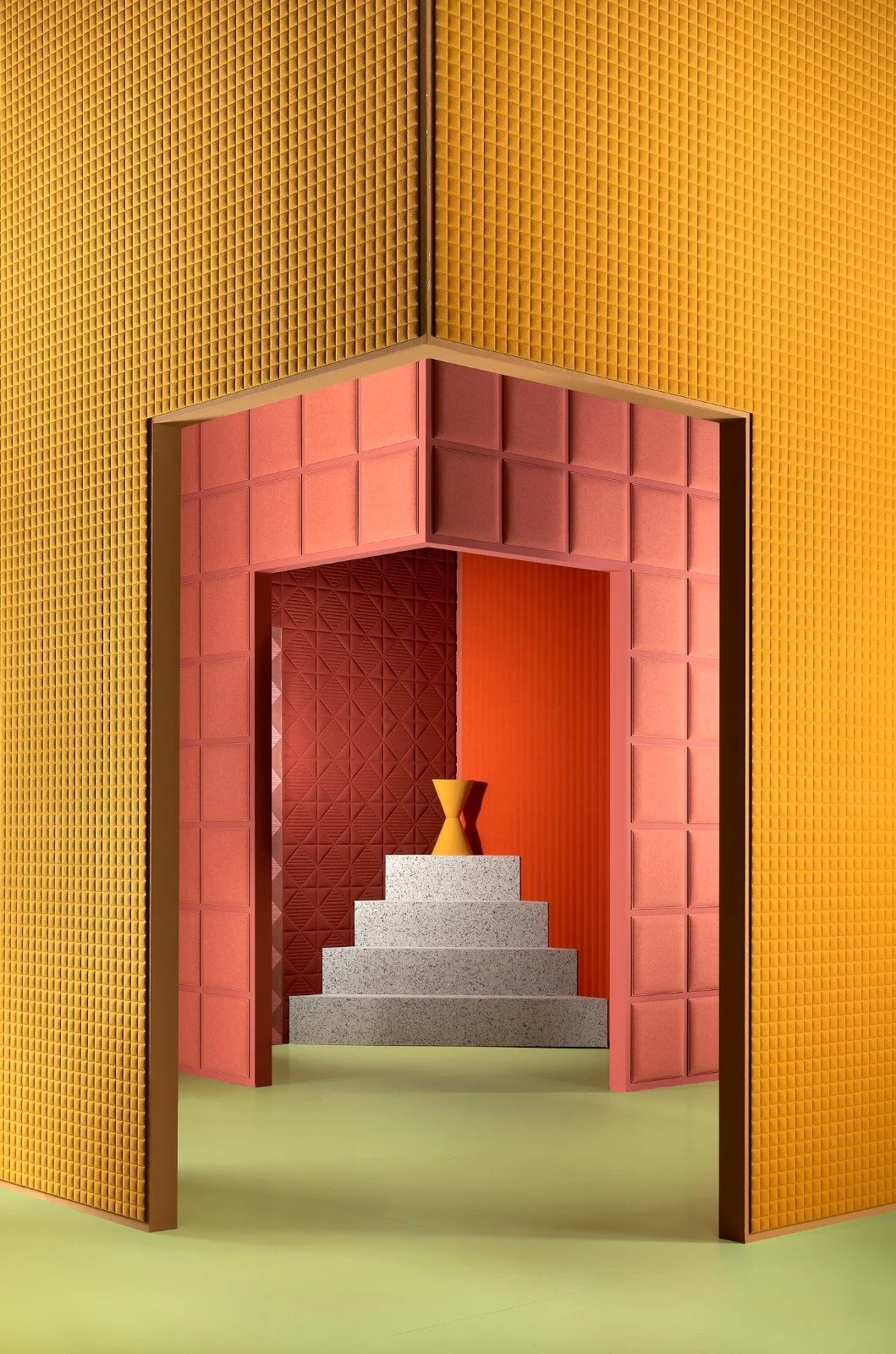 An art installation featuring a pink geometric structure with steps, a yellow vase on top, framed by yellow and orange textured walls.