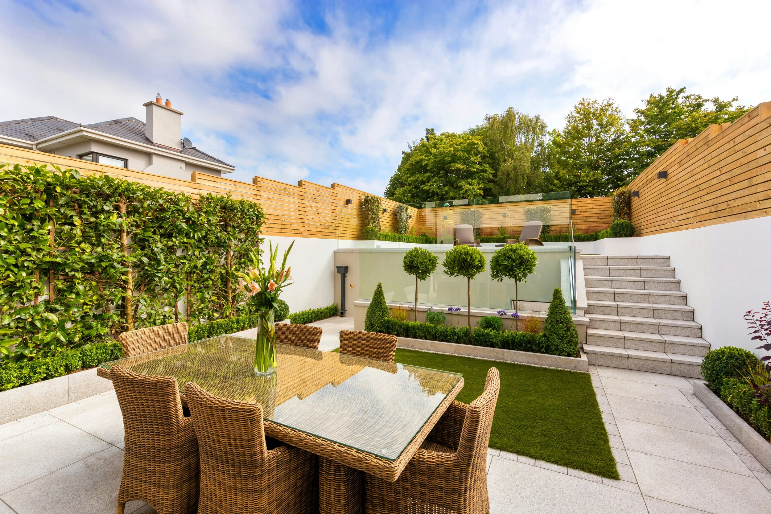 A modern backyard patio with a glass-topped dining table and six wicker chairs, a small lawn, and a garden area with plants, shrubs, and trees, enclosed by wooden fencing and white walls with steps leading up to a raised seating area.