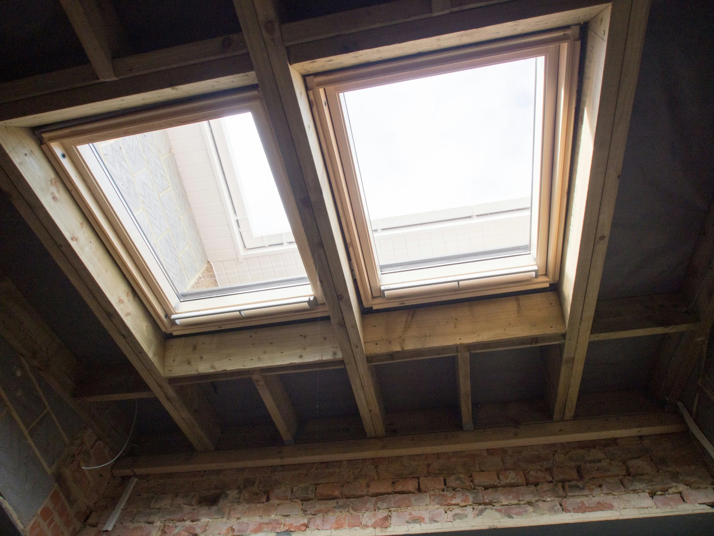 Two large skylight windows installed in a wooden ceiling framework, with brick wall below.