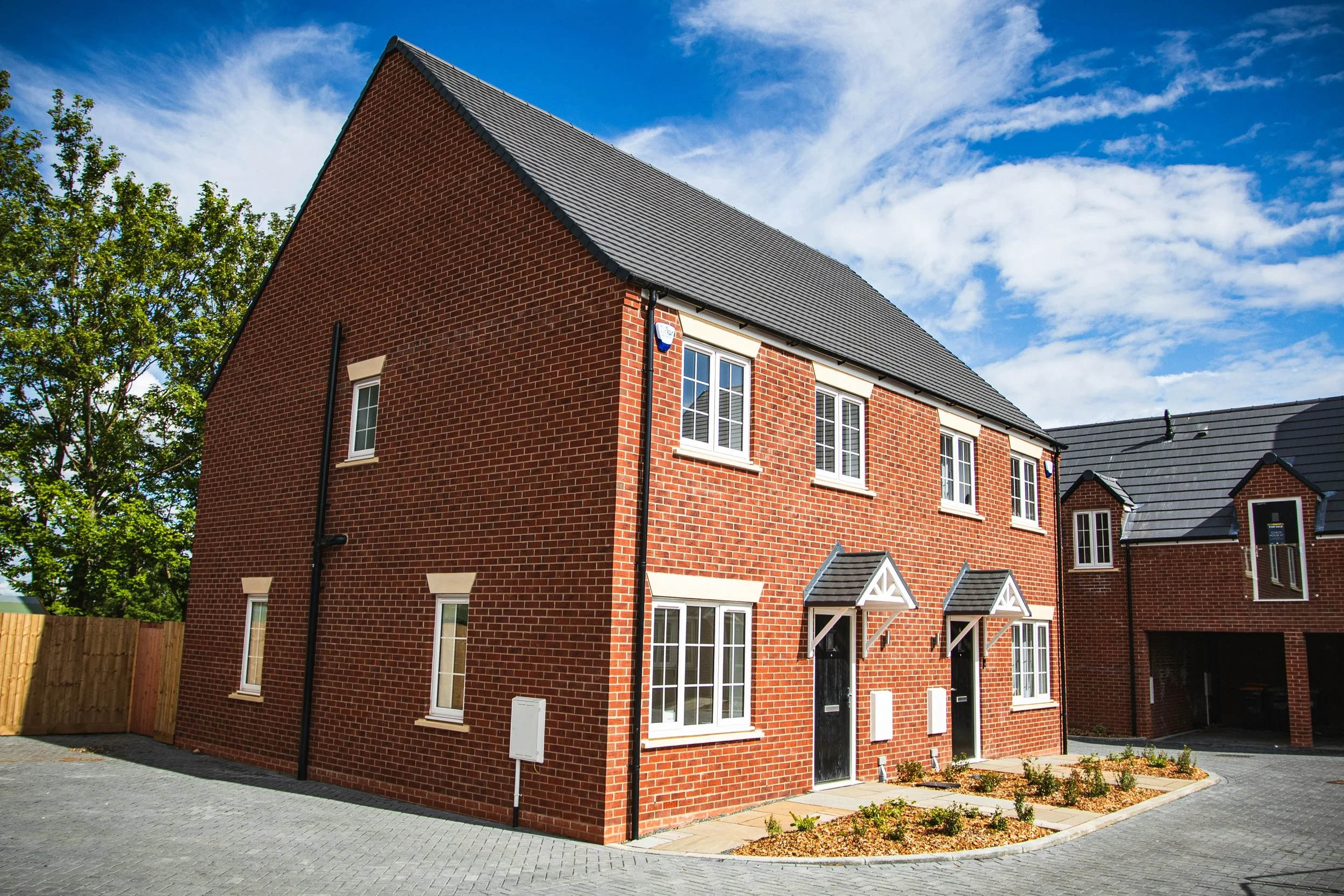 Newly built red brick townhouse with black front doors, small window awnings, and a landscaped front yard, under a partly cloudy blue sky.