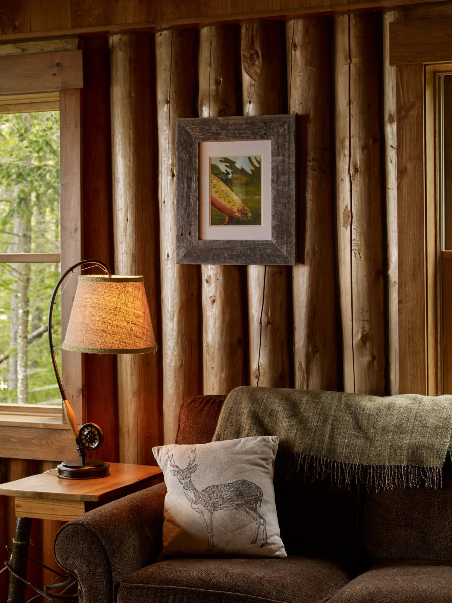 Living room with wooden paneled wall, a framed animal picture, a soft armchair with a deer-printed pillow and threw blanket, a wooden side table, a table lamp, and a window showing trees outside.