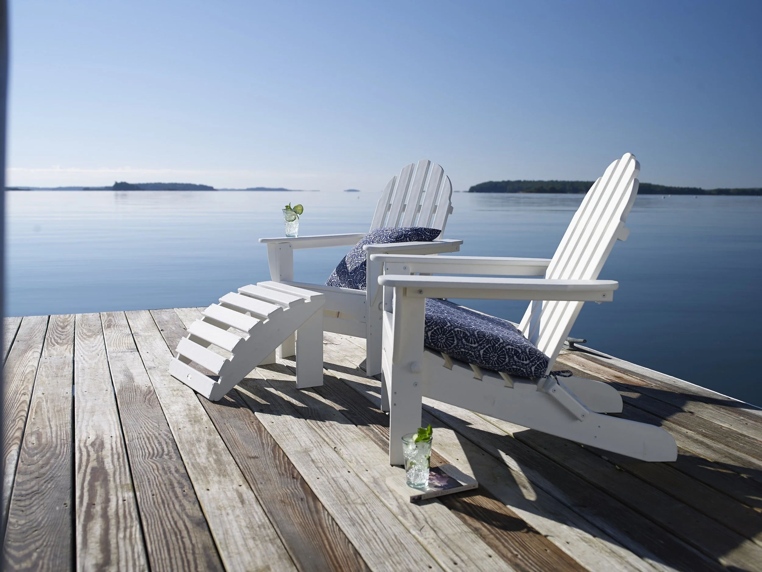 Two white Adirondack chairs on a wooden dock overlooking a calm body of water with distant islands under a clear blue sky, with two glasses of mojitos on the dock.