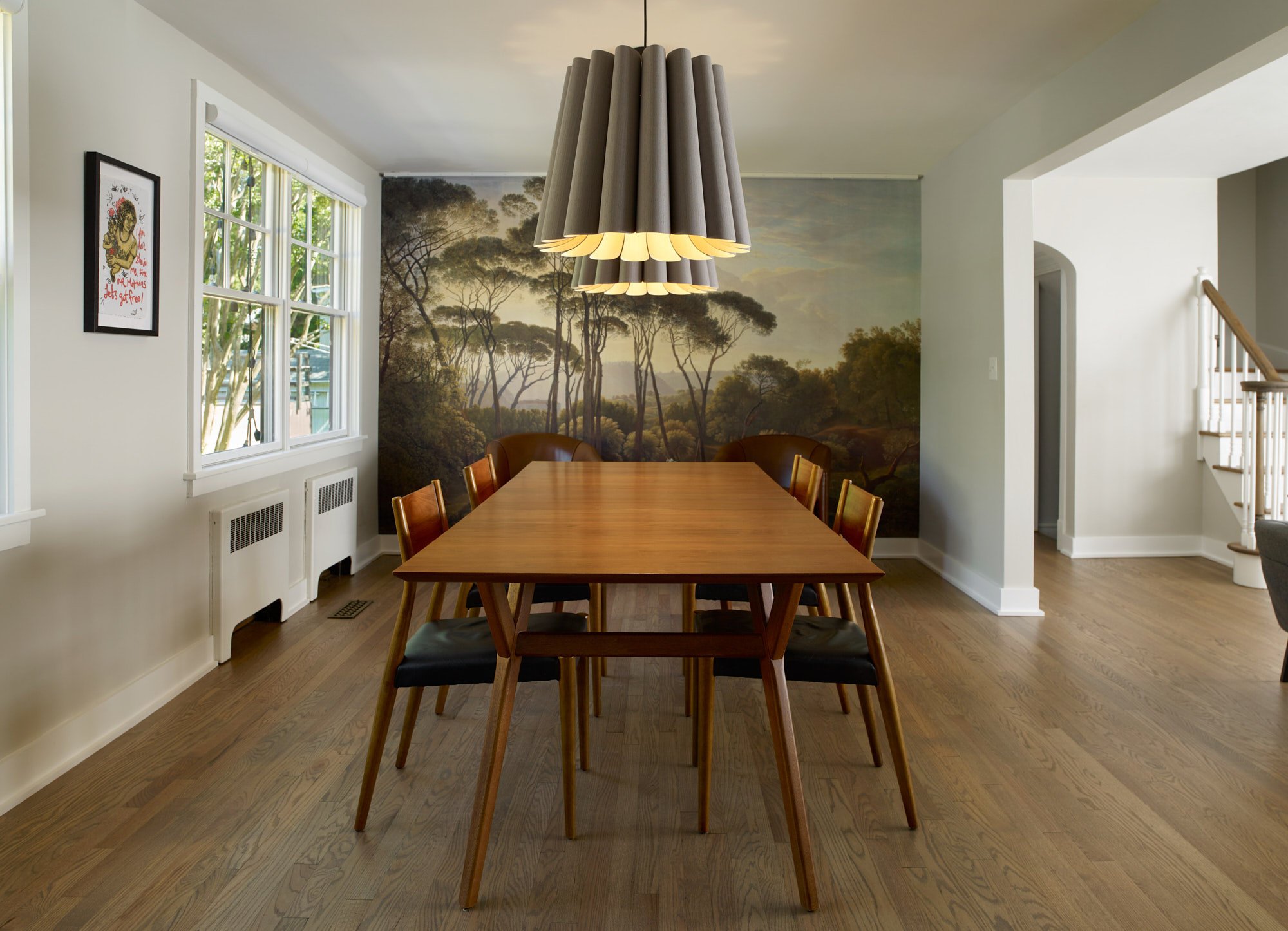 A wooden dining table with six chairs in a room with a scenic forest mural on the wall, a window showing green trees, and a modern pendant light fixture hanging above the table.