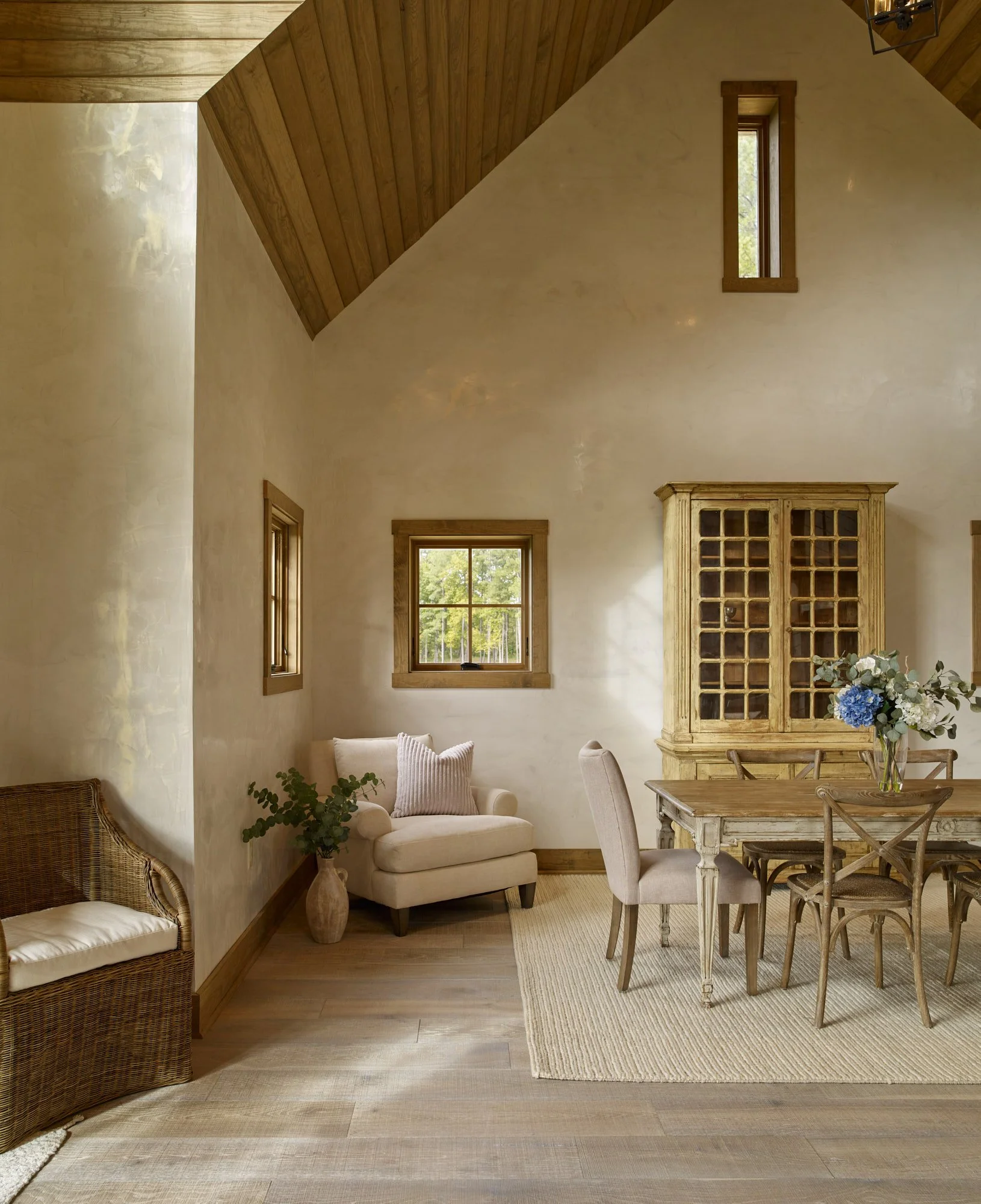 Living room with beige walls, wooden ceiling, windows with blinds, a white armchair with a pillow, wooden dining table with chairs, decorative flowers, and a wooden cabinet.