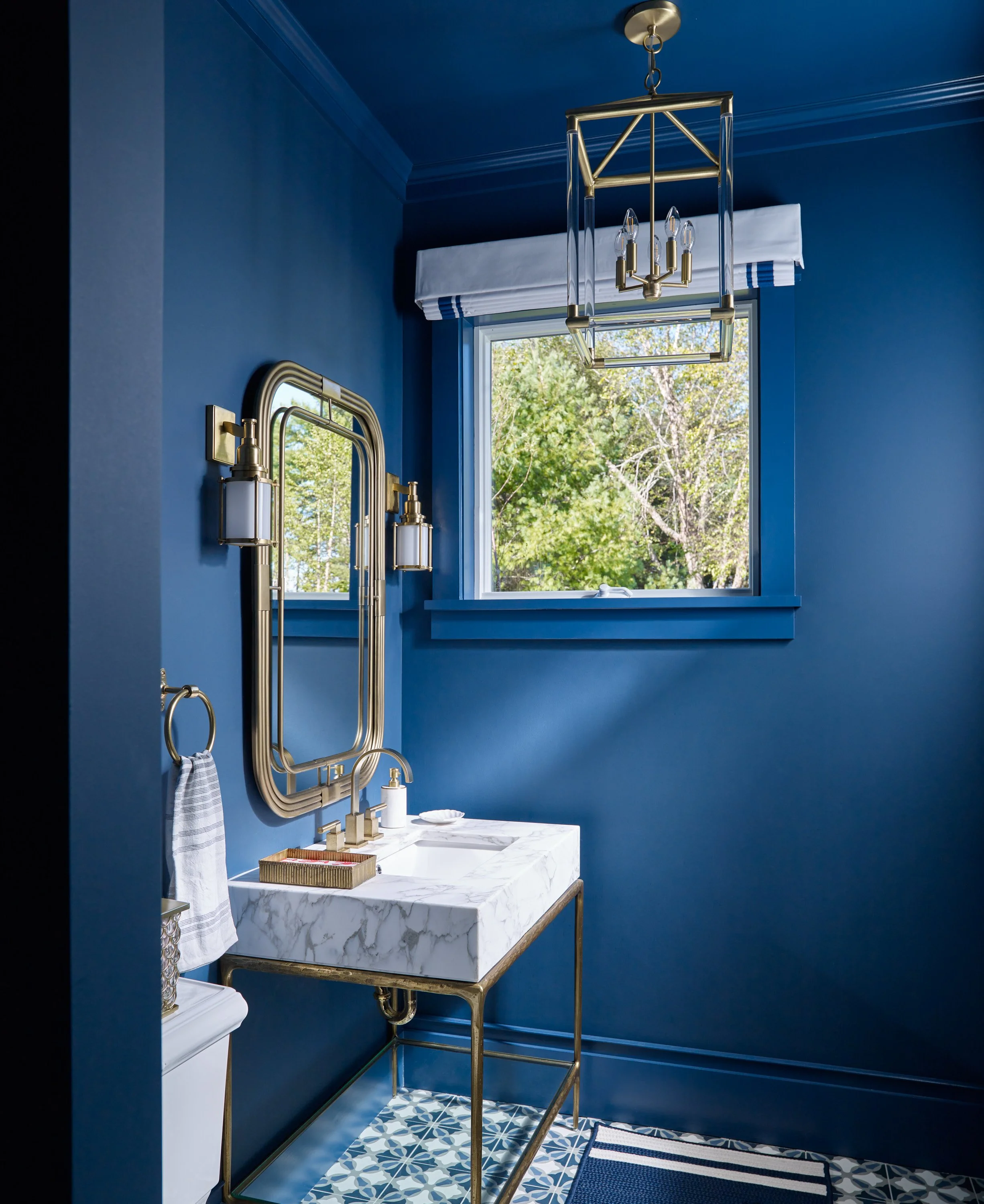 A modern bathroom with vibrant blue walls, a white marble vanity with gold accents, and a geometric patterned floor. There is a window with a white valance, a gold and glass chandelier hanging from the ceiling, and two matching gold and glass wall sc