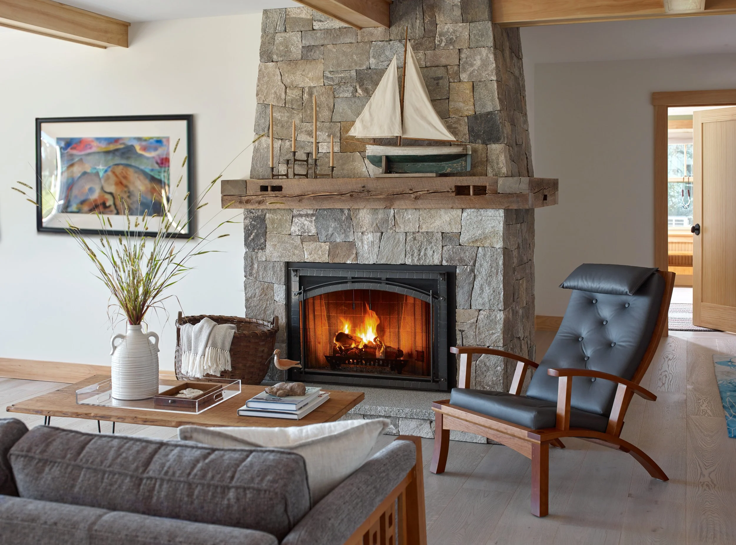 Living room with a stone fireplace, a small sailing boat decoration above the mantle, a candelabra with lit candles, a potted plant, a wooden coffee table with books and decorative items, and a comfortable armchair near the fireplace. Open doorway le