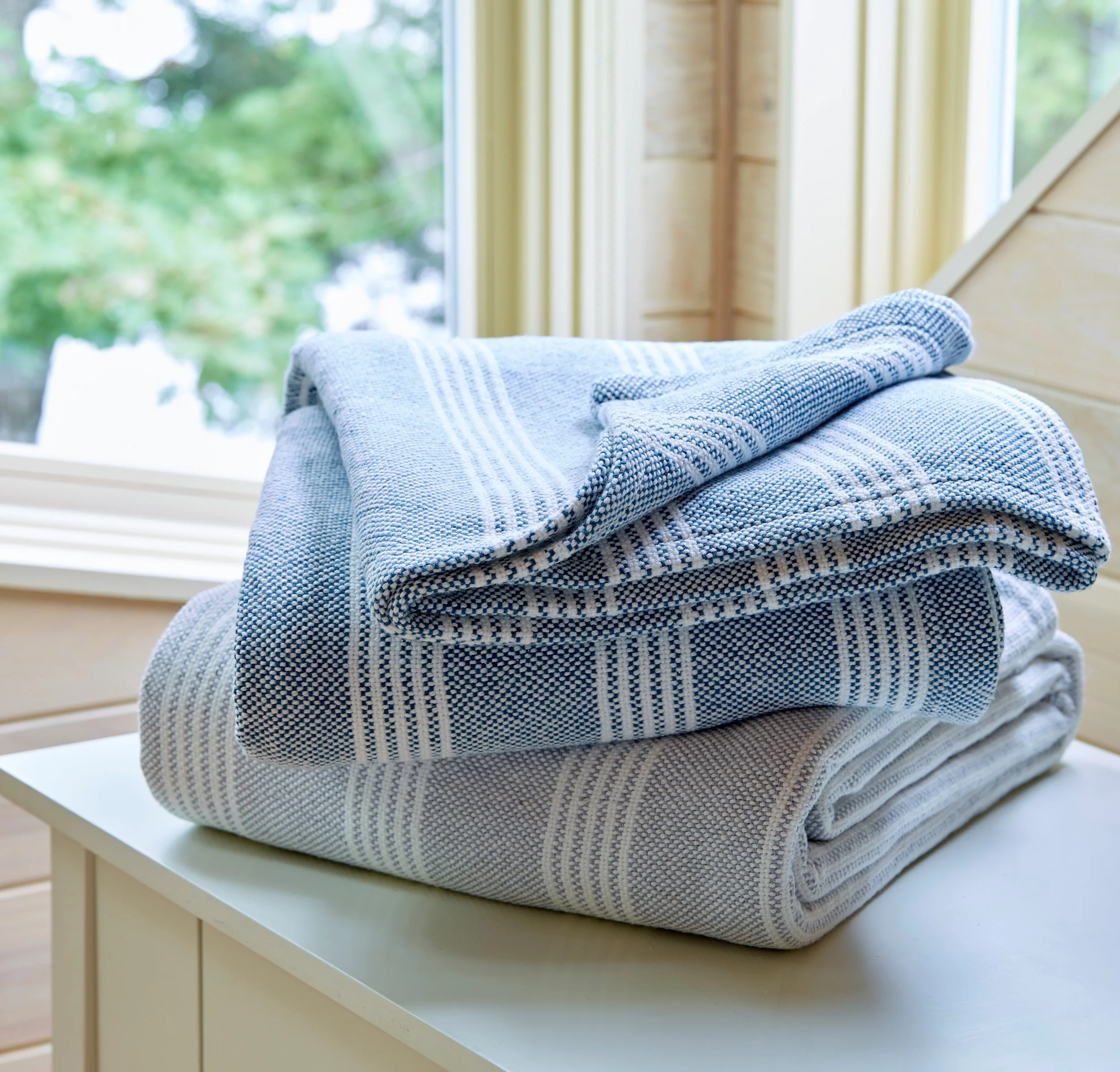Stacked blue and white striped towels on a light-colored table with a window and greenery in the background.