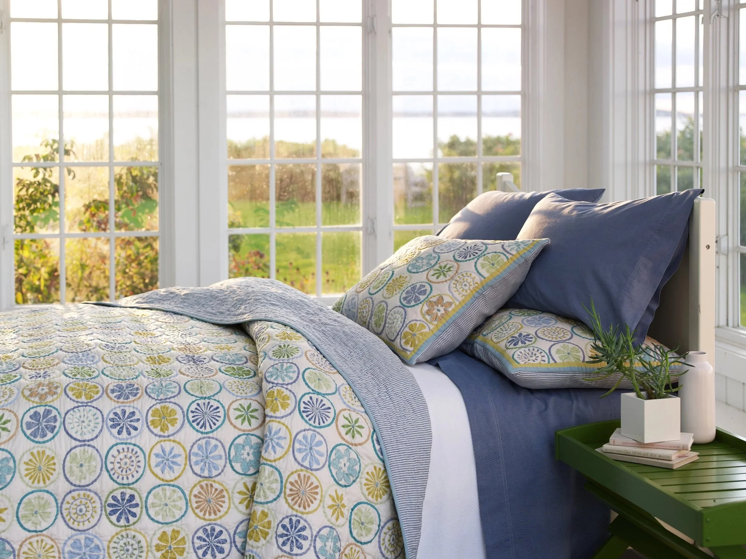 Sunlit bedroom with large windows showing greenery outside, bed with colorful floral patterned quilt and dark blue pillows, green side table with books and a potted plant.