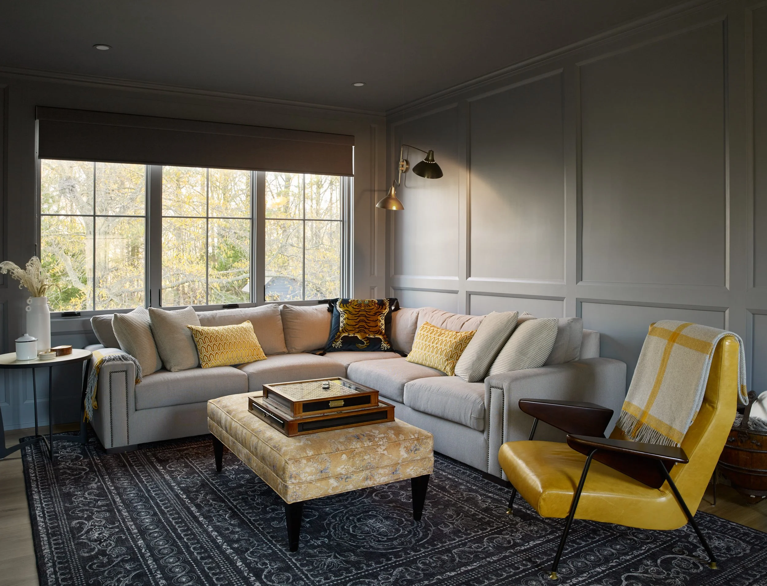 Living room with beige sofa, yellow and patterned cushions, glass-topped side table, upholstered ottoman, black and yellow armchair, large window with view of trees, wall sconce lighting, dark patterned rug.