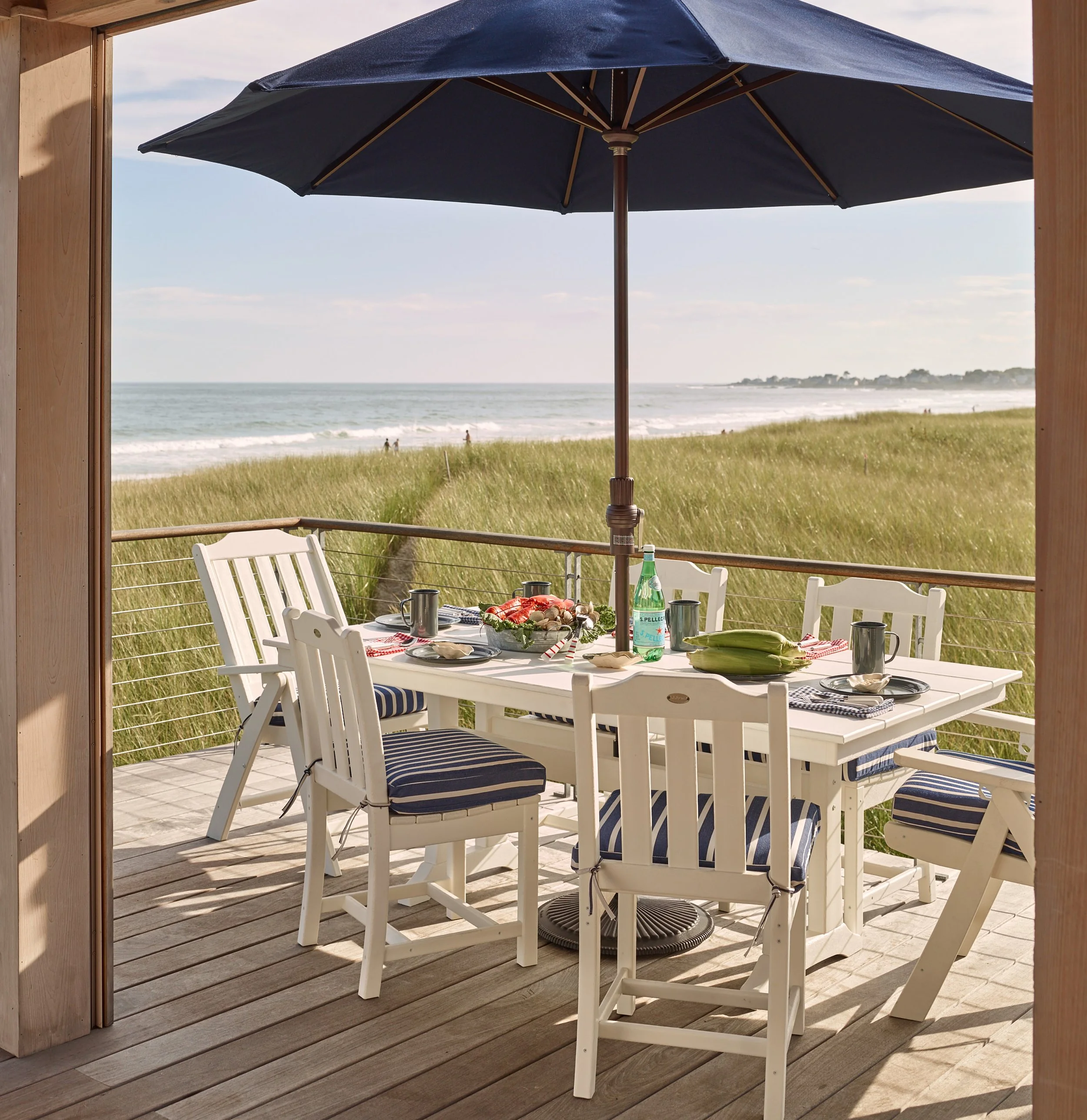 A dining table set on a wooden deck overlooking a beach, with an umbrella, food, and drinks, framed by a wood structure.