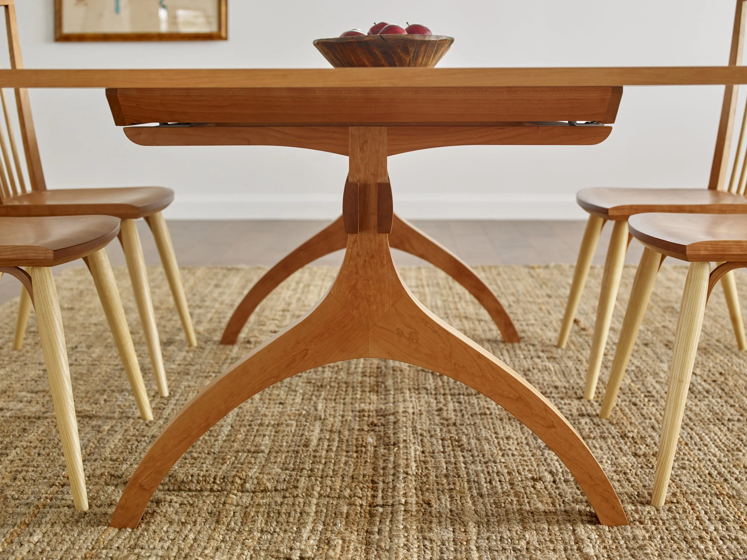 A wooden dining table with a bowl of red apples on top, surrounded by wooden chairs with curved backs, on a textured beige rug in a room with white walls and a framed picture in the background.