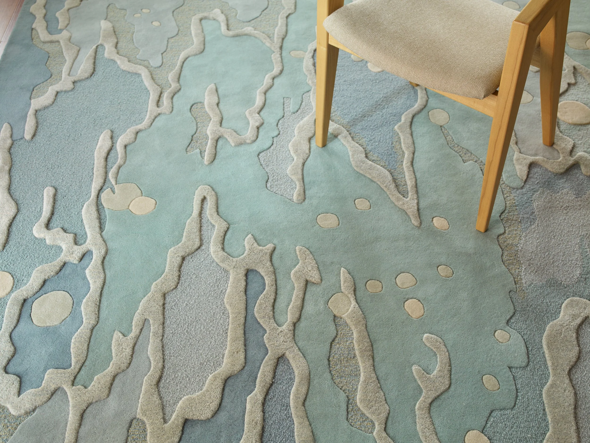 Close-up of a light blue area rug with irregular, abstract cream-colored patterns and a wooden chair with a beige upholstered seat.