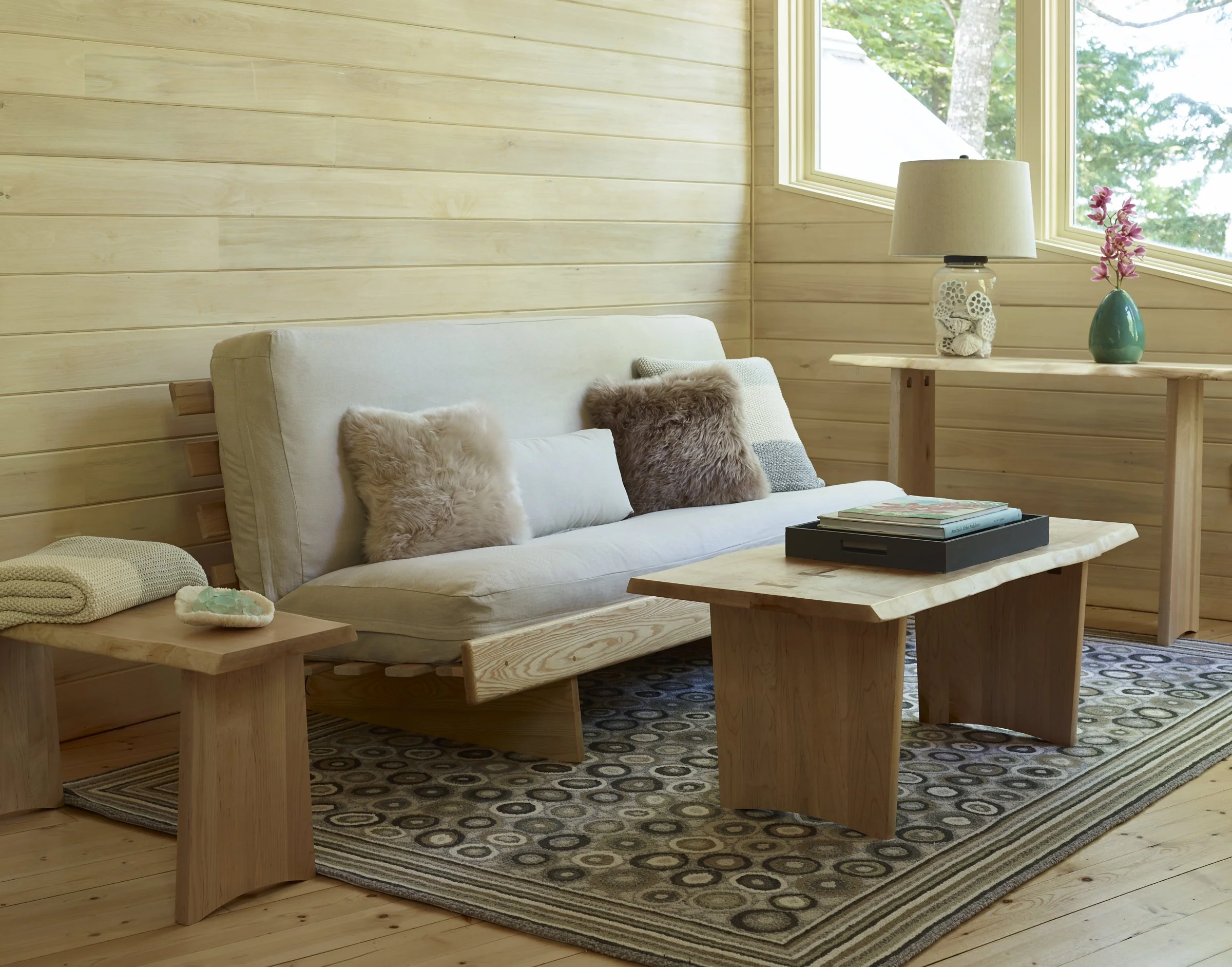A cozy living room with wooden walls and large windows showing green trees outside. It features a white sofa with fluffy pillows, a wooden coffee table with books, and a side table with a lamp and a vase of pink flowers.