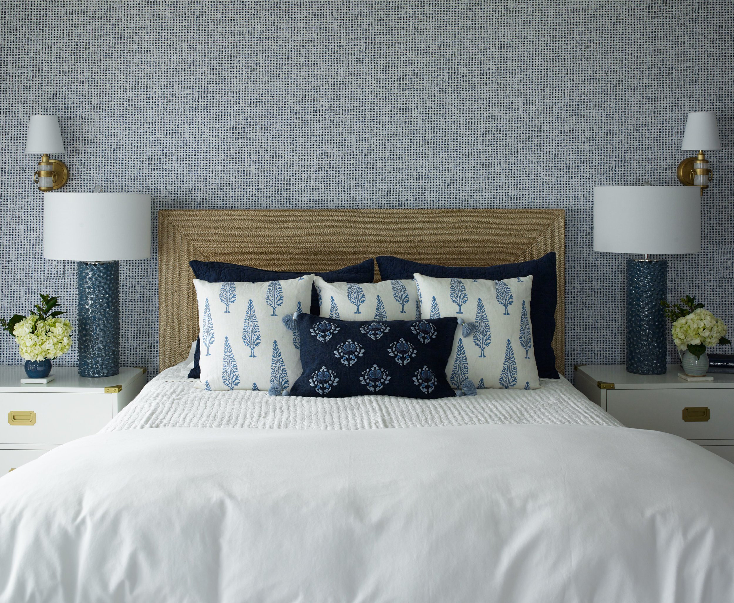 A neatly made bed with white bedding and navy blue pillows, flanked by two white nightstands with blue and gold lamps, against a blue patterned wall.