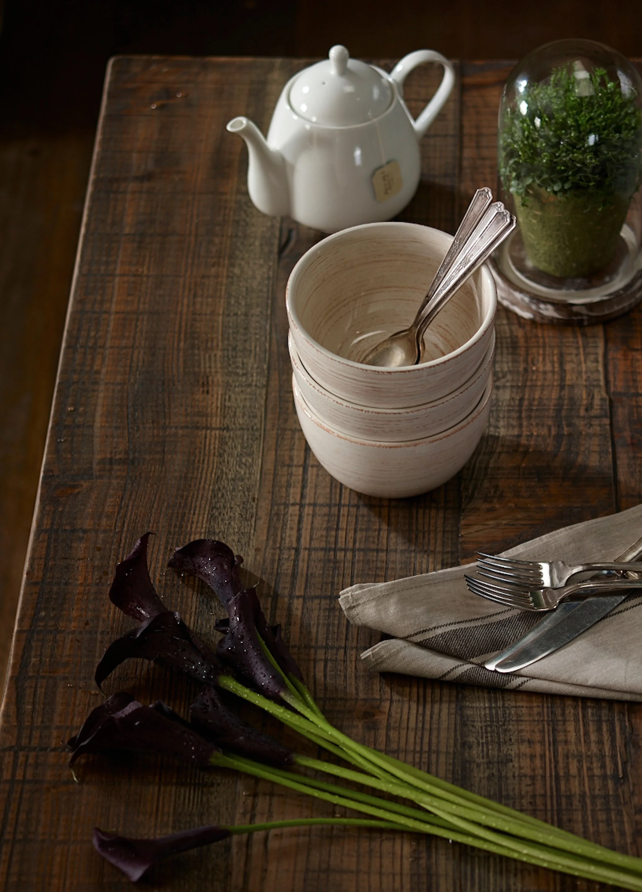 A rustic wooden table set for a meal with a white teapot, a stack of three ceramics bowls with a spoon inside, a glass dome covering a small potted plant, and a bunch of dark purple and green leafy vegetables on the side, with a fork and knife wrappe