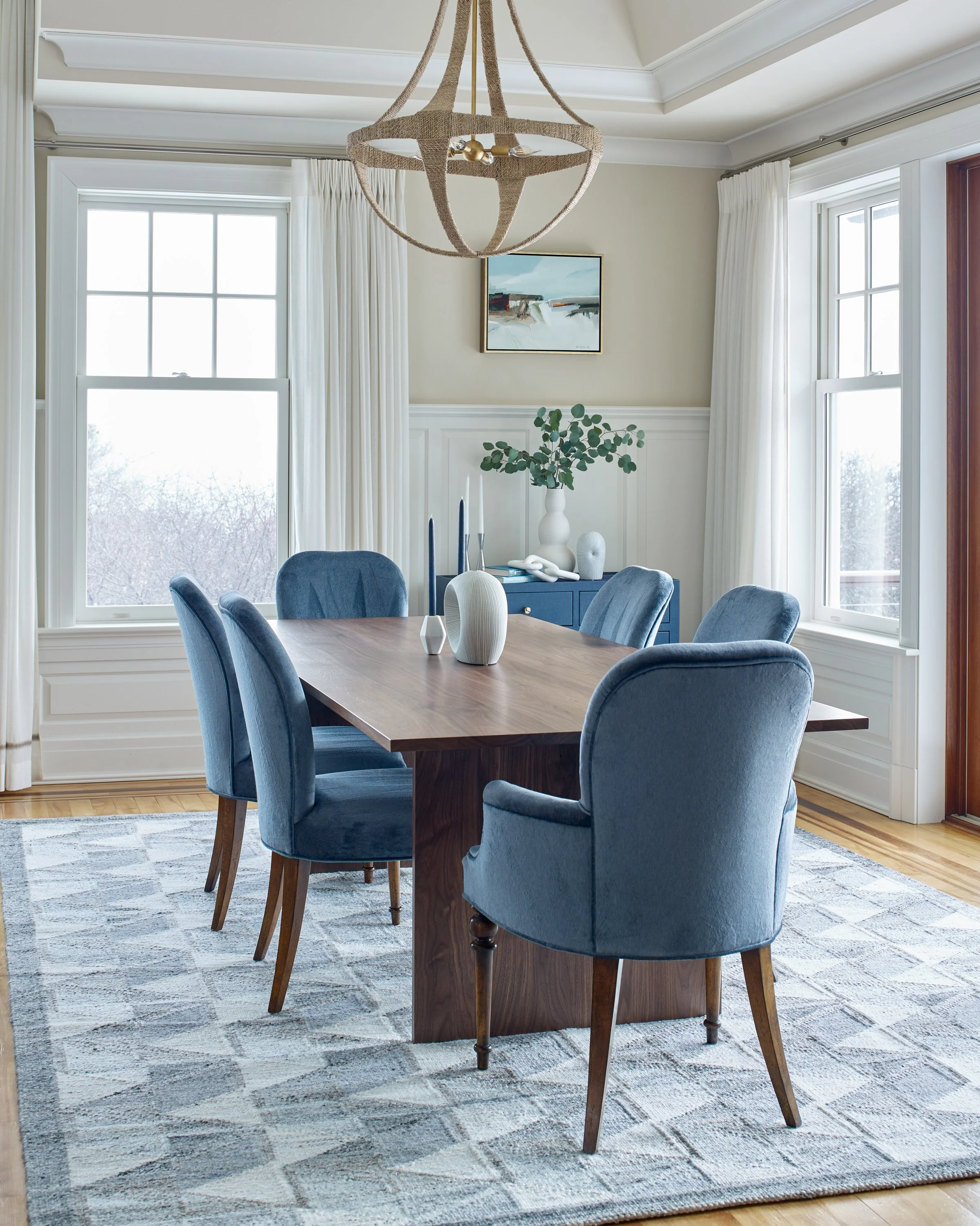 A dining room with a wooden table, surrounded by six blue upholstered chairs. On the table, there are vases and candles. In the background, a blue sideboard features decorative objects and a large green leafy plant. Three large windows with white cur