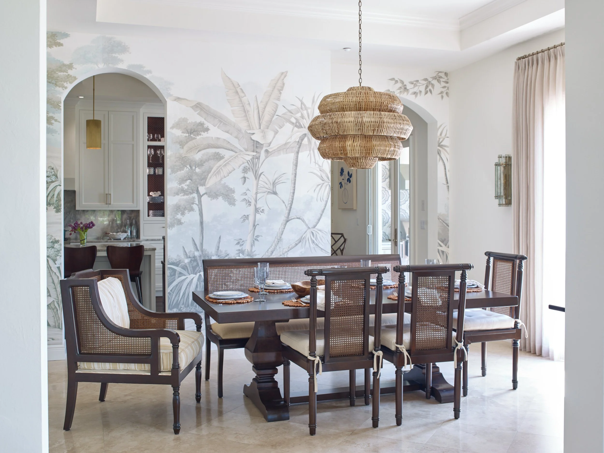 A dining room area with a dark wood table set with plates, cloth napkins, and clear glasses, surrounded by chairs with woven cane backs and cream-colored cushions. A woven pendant light hangs above the table. The background features a wall with a bot